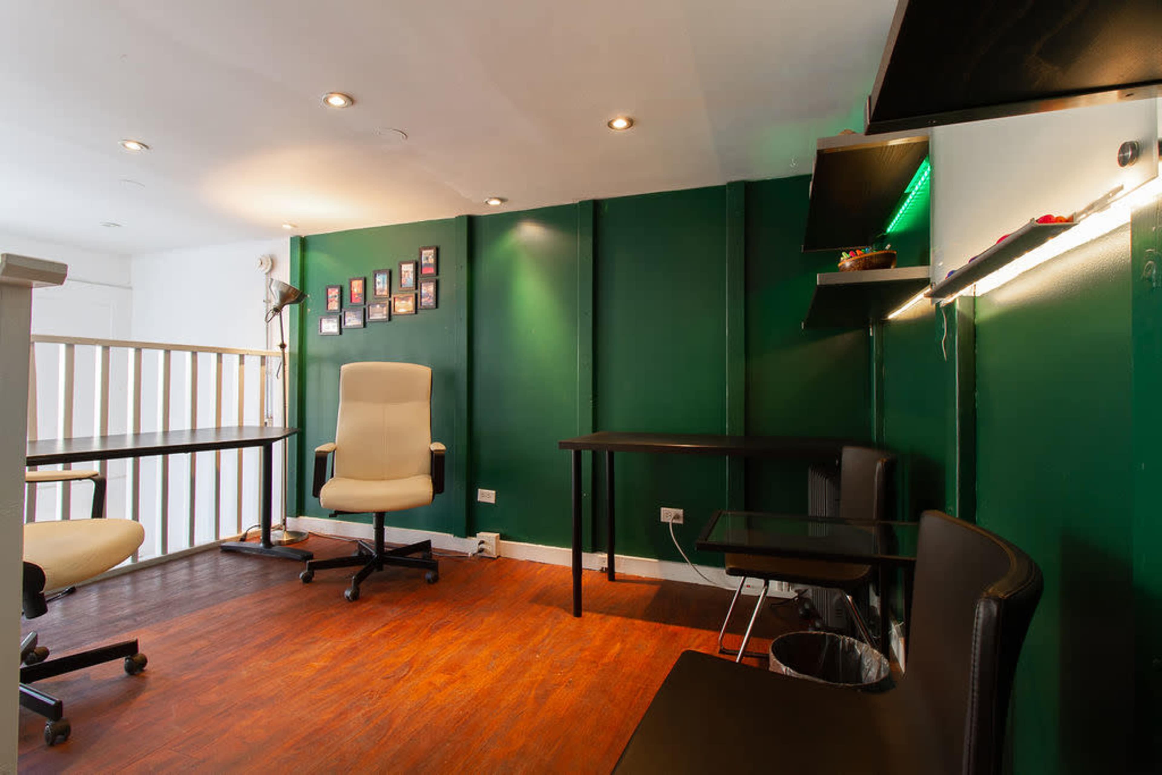 The image shows a small office space with green walls, wooden flooring, a desk, an office chair, and a wall displaying several framed photographs.