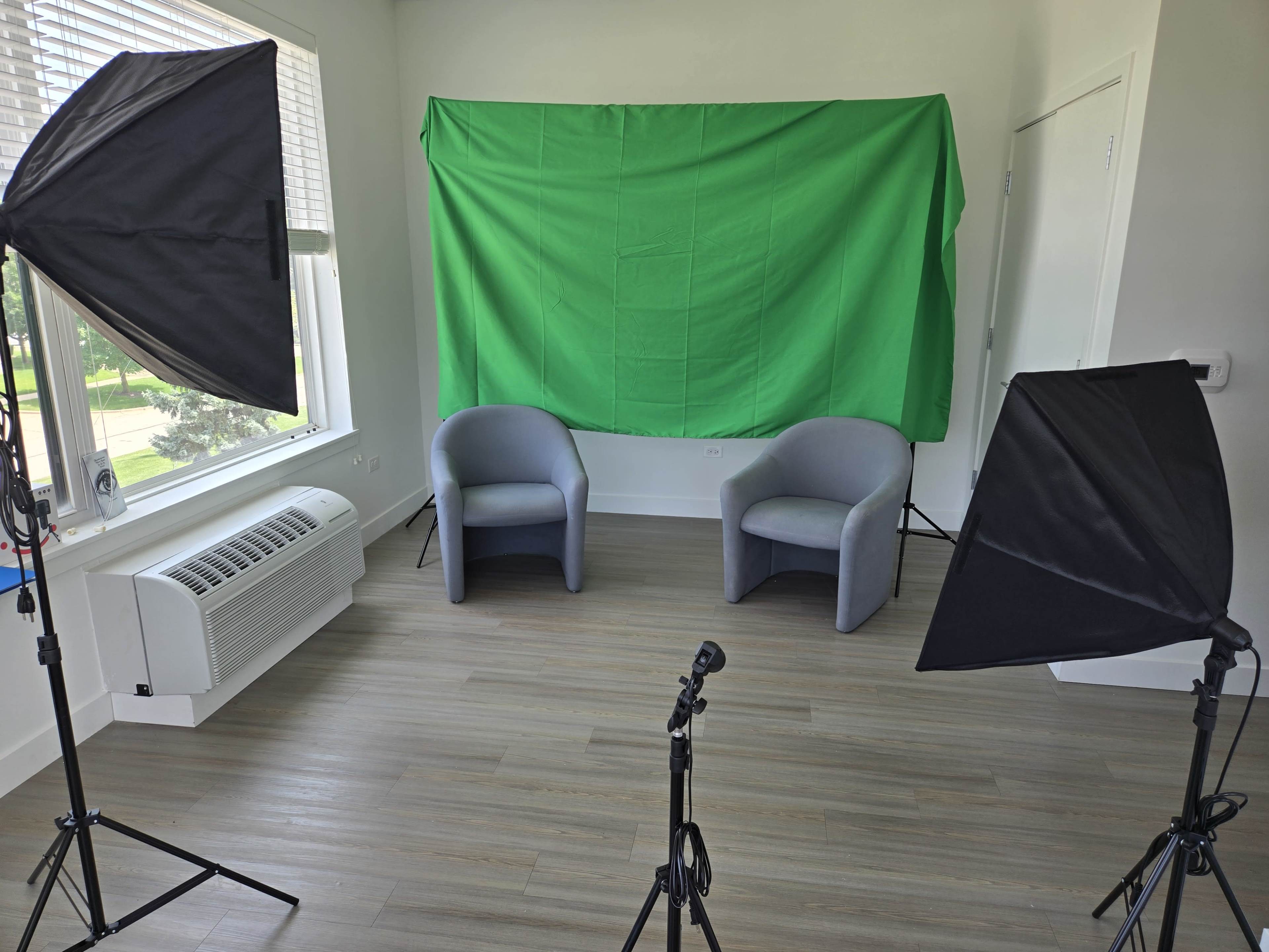 The image shows a brightly lit room with two gray chairs in front of a green backdrop, flanked by two photography softboxes on tripods.