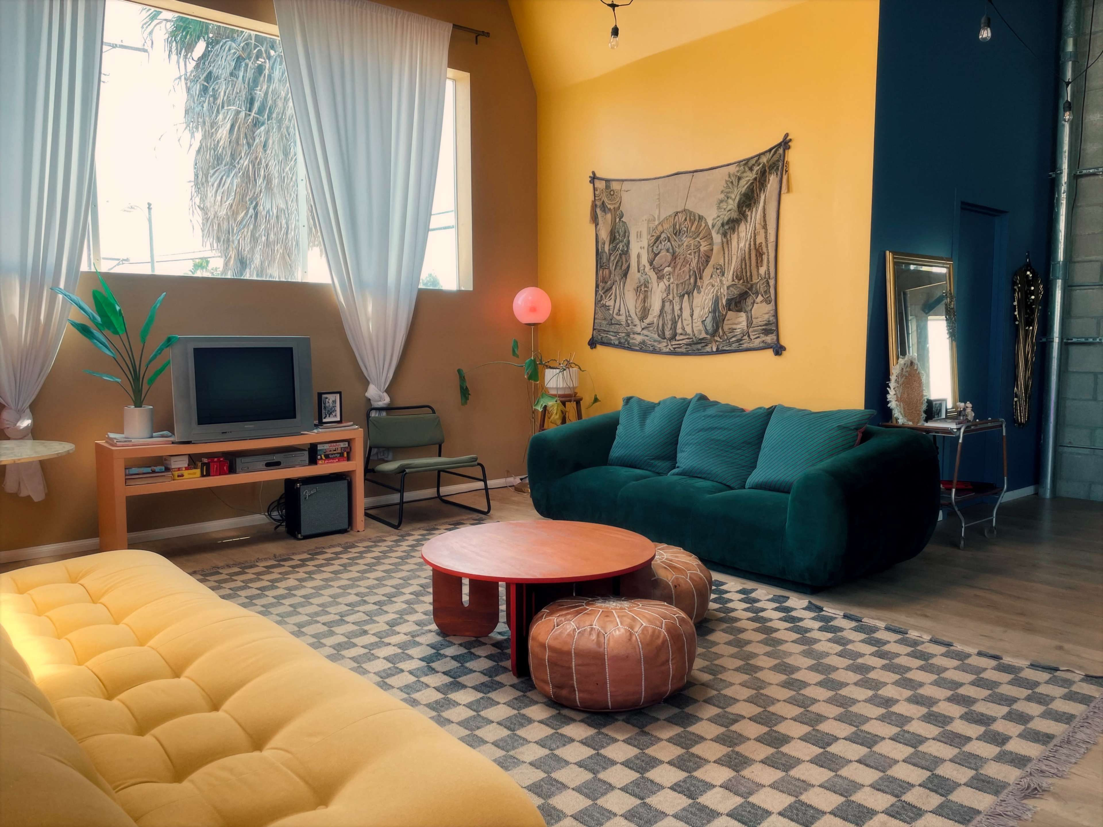 The image shows a modern living room with a yellow wall, a green sofa, a yellow couch, a round coffee table, and large windows with draped curtains.