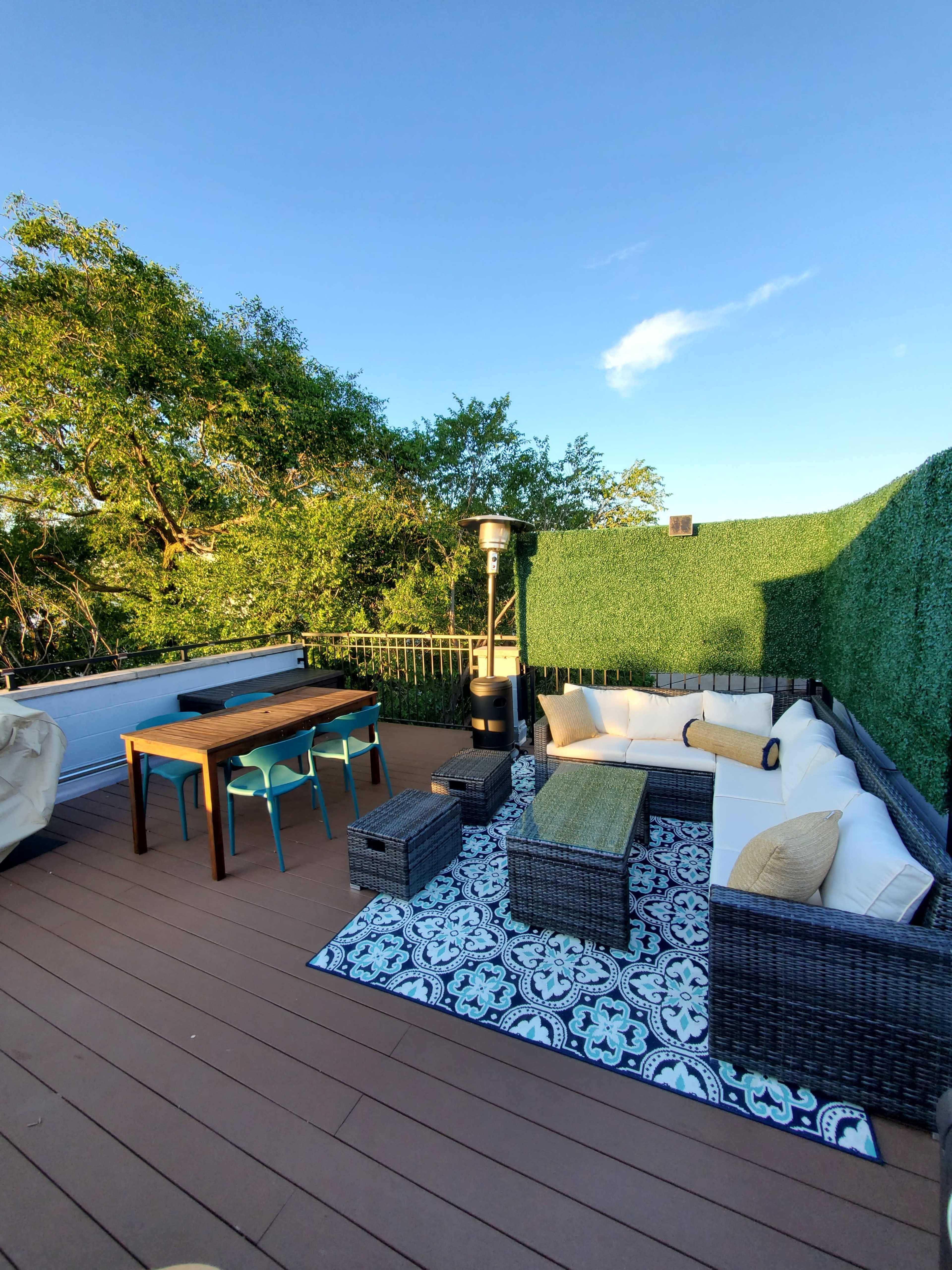 A rooftop terrace featuring a seating area with white couches, a table with blue chairs, and a patterned rug surrounded by greenery.