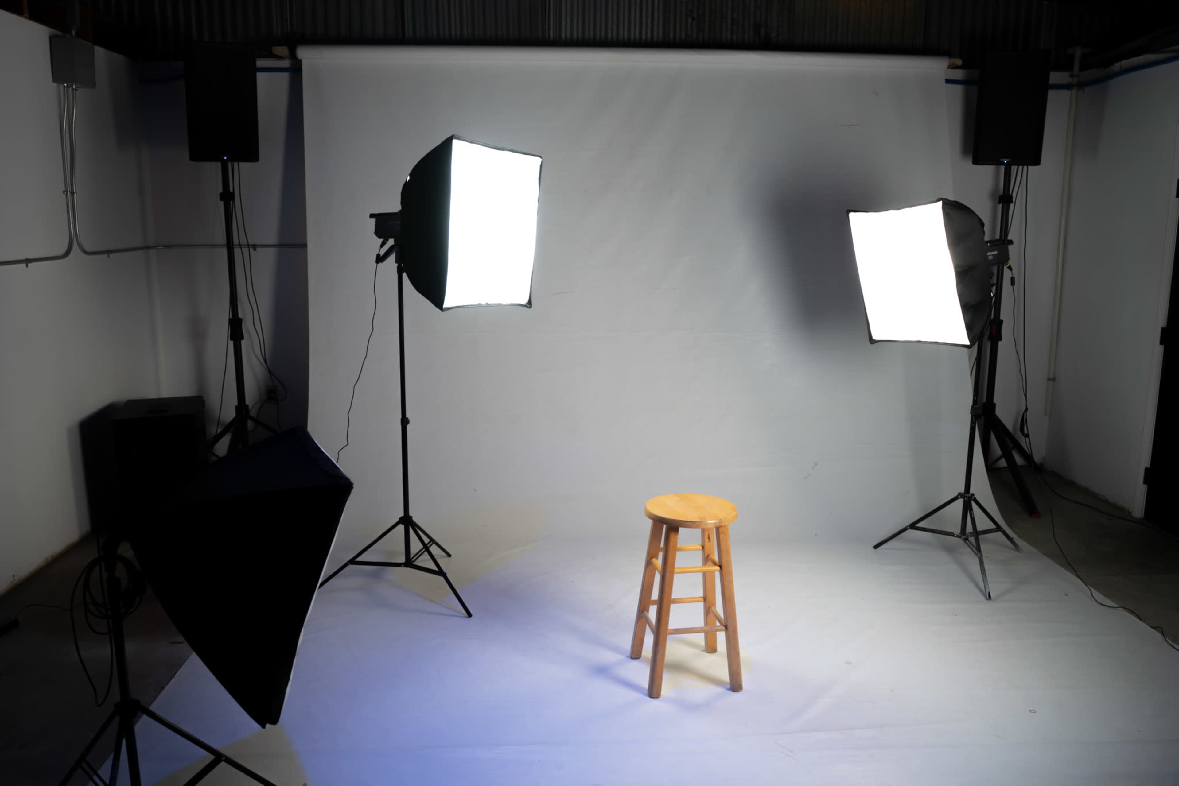 A wooden stool is positioned in front of a plain white backdrop, illuminated by two large softbox lights and flanked by speakers.
