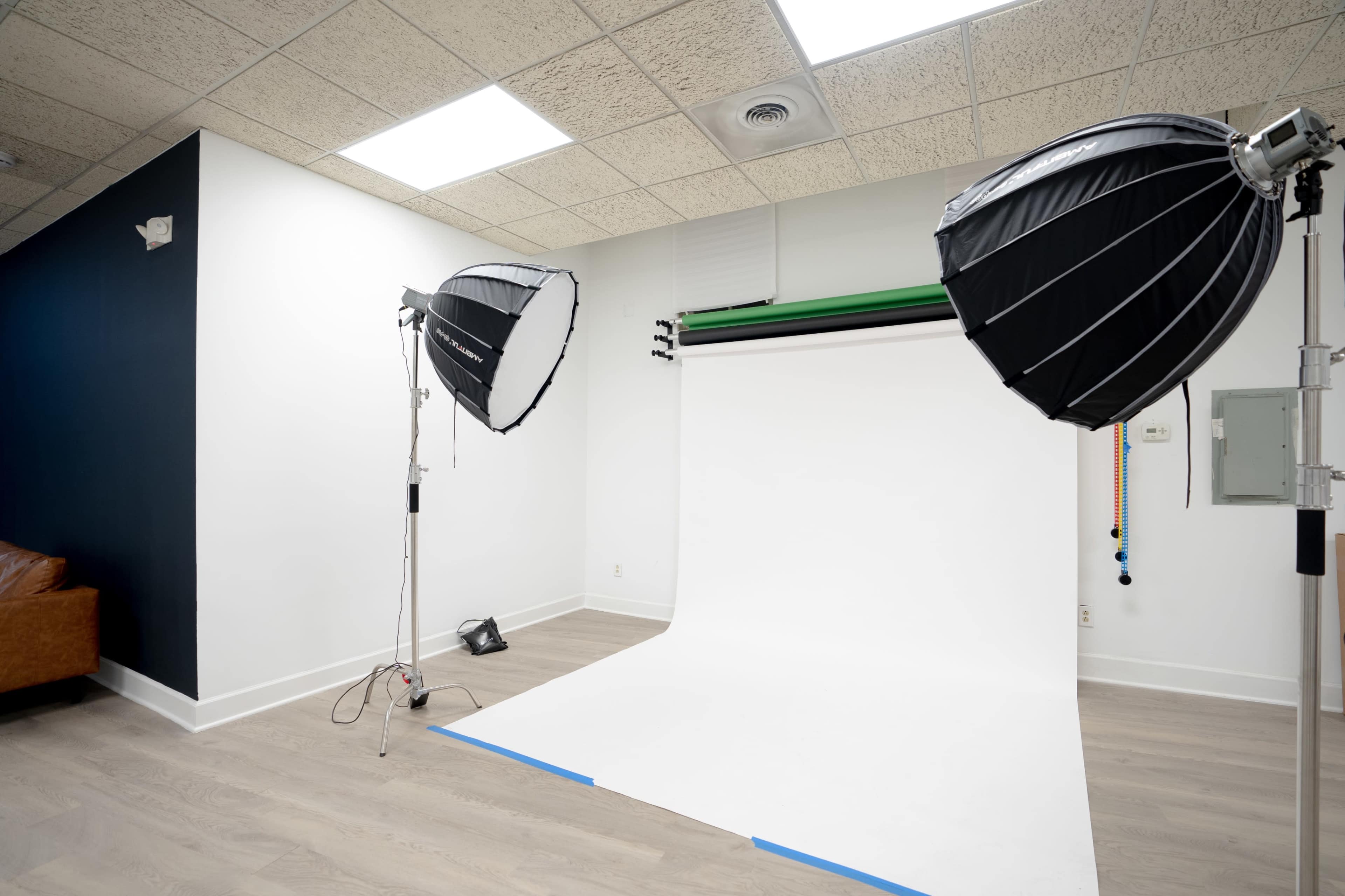 The image shows a photography studio with a white backdrop, two softbox lights, and a brown couch in the corner.