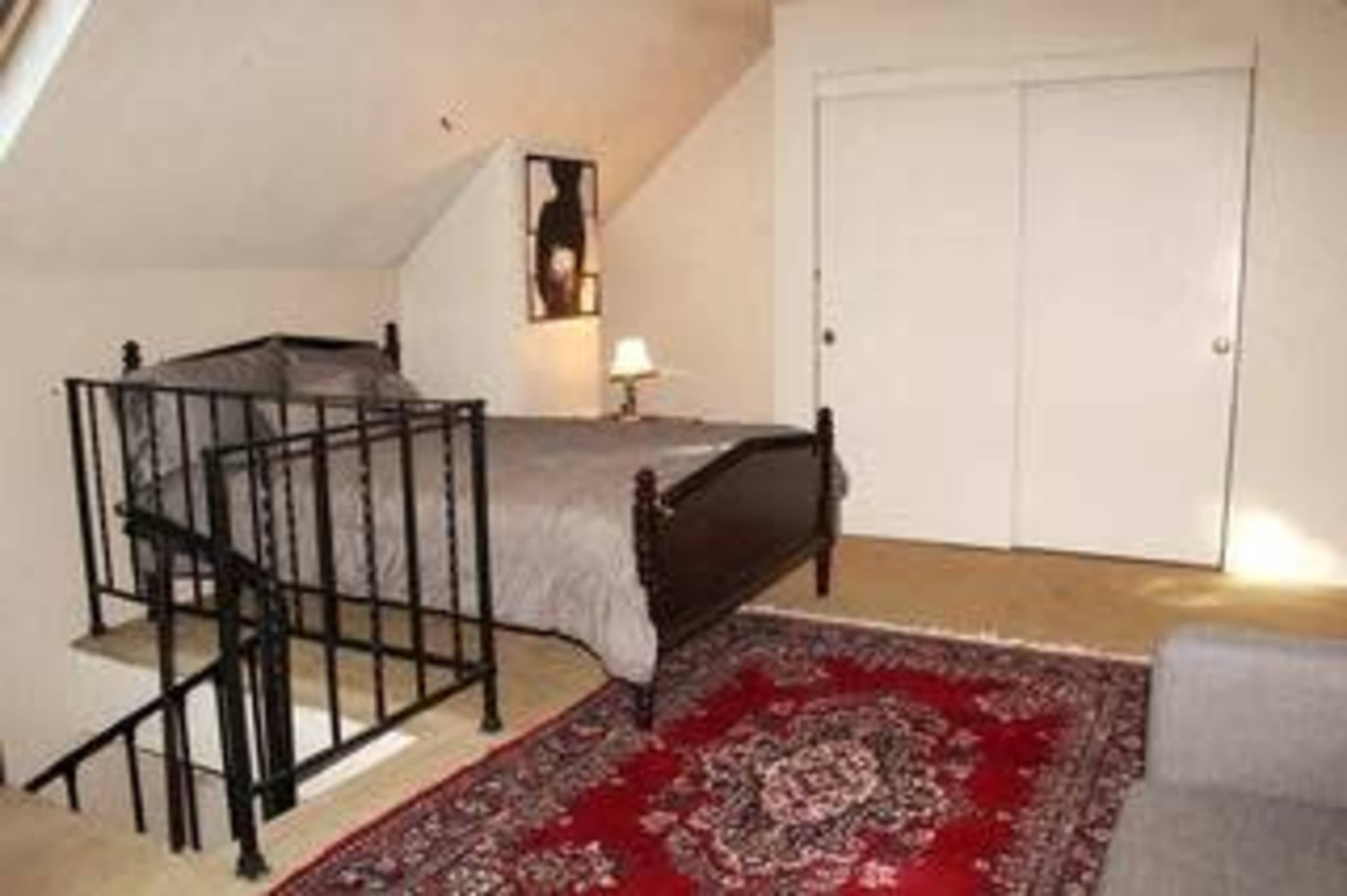 A cozy attic bedroom features a bed with a gray comforter, a small lamp on a bedside table, and a red patterned rug on a beige carpet.