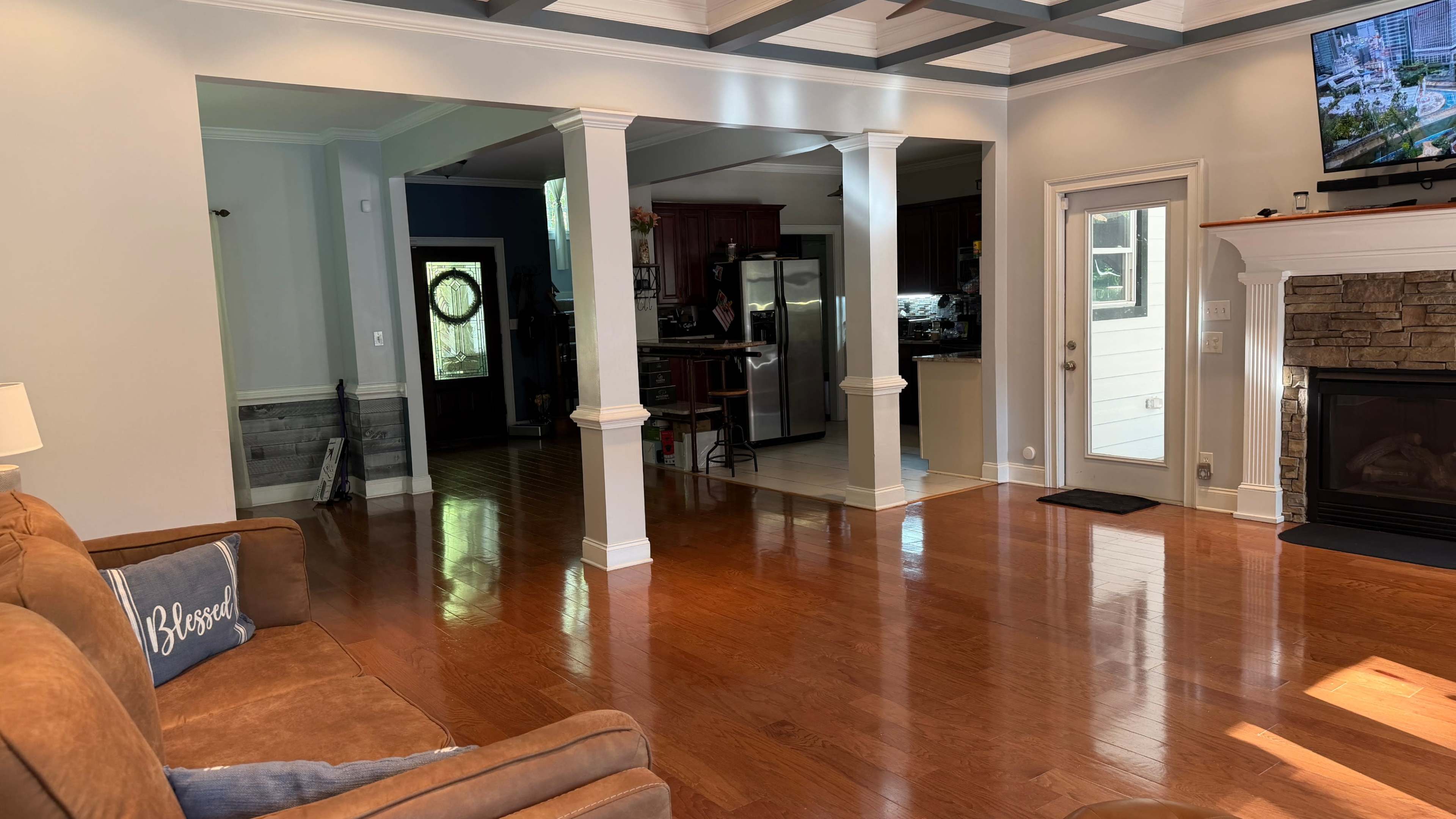 The image shows a spacious living room with polished wooden floors, a brown sofa, and a view of a kitchen area through an archway.