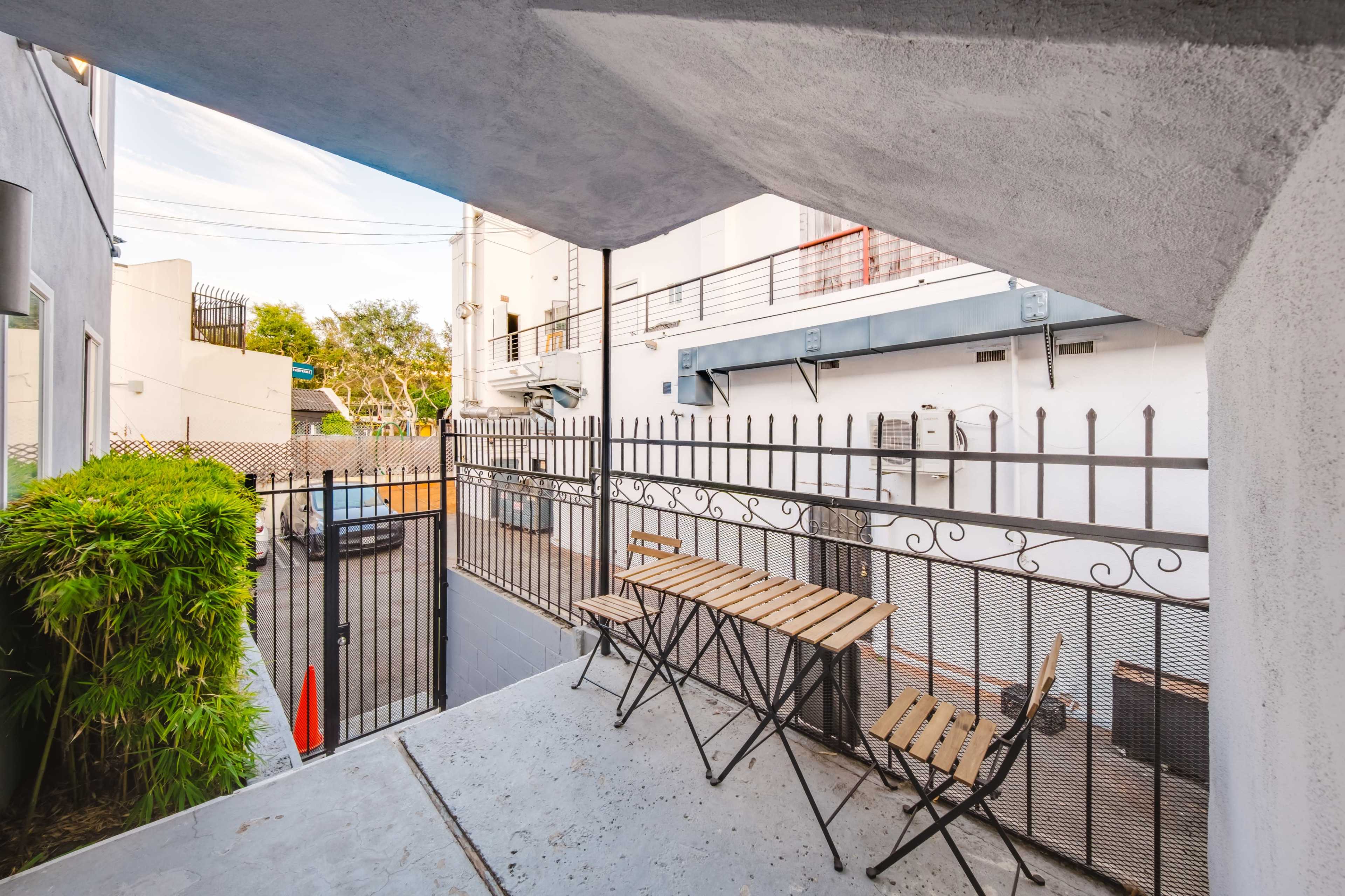 The image shows a small outdoor patio area with a metal fence, a table, and chairs, situated between two buildings.