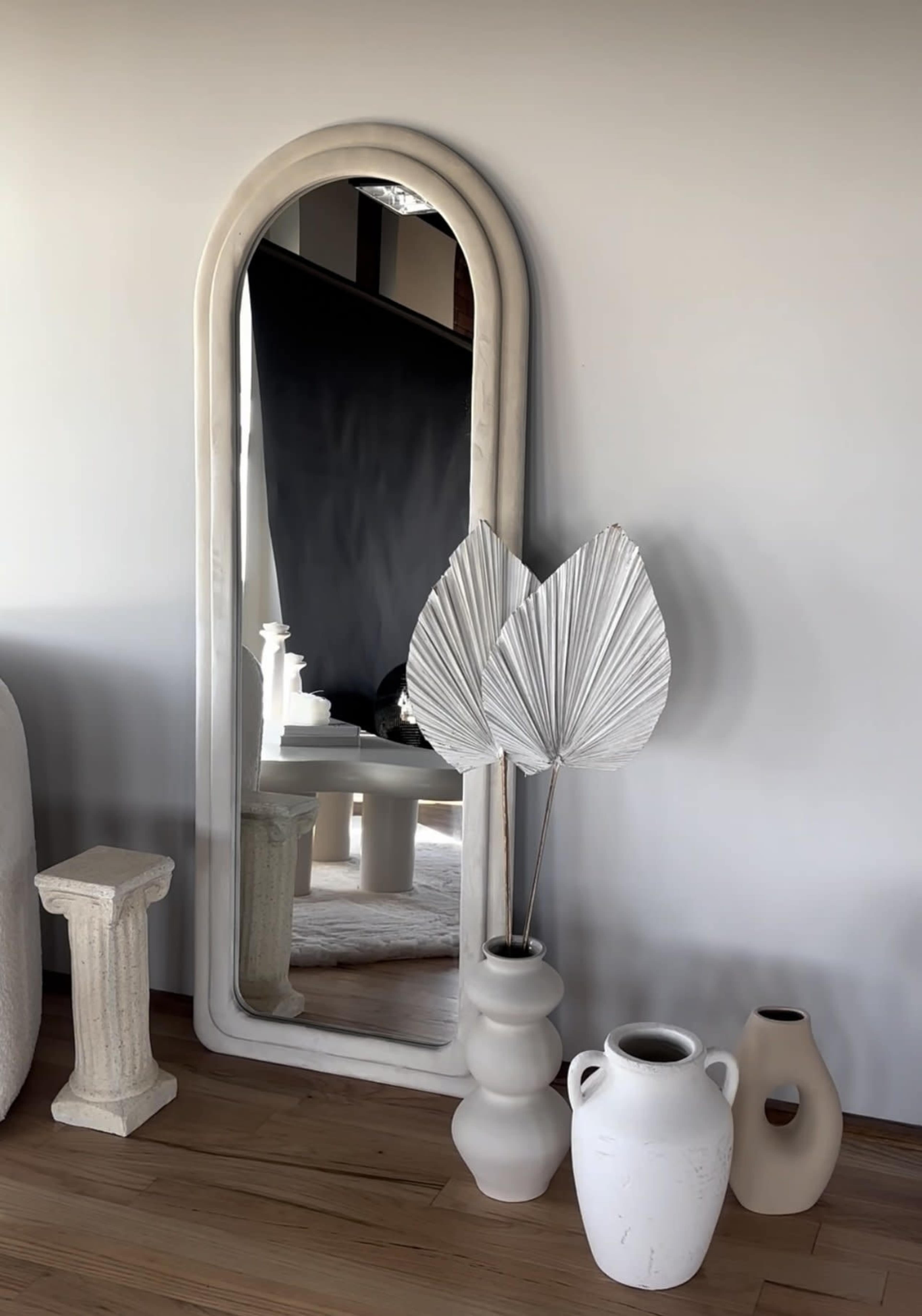 A tall, arched mirror stands against a wall, surrounded by decorative vases and a large, dried plant.