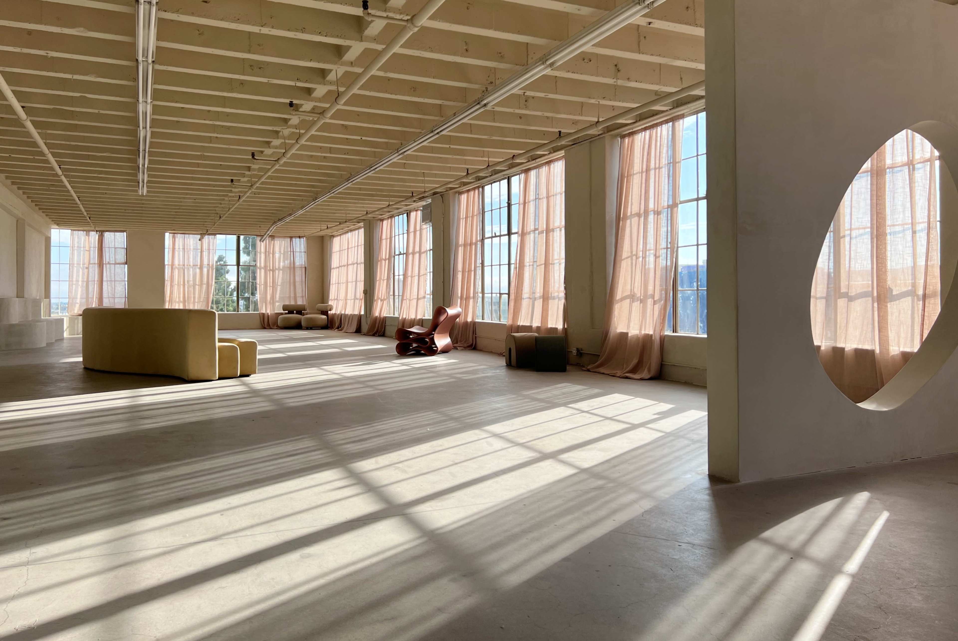 The image shows a spacious, well-lit room with large windows, sheer curtains, and minimal furniture along the walls.