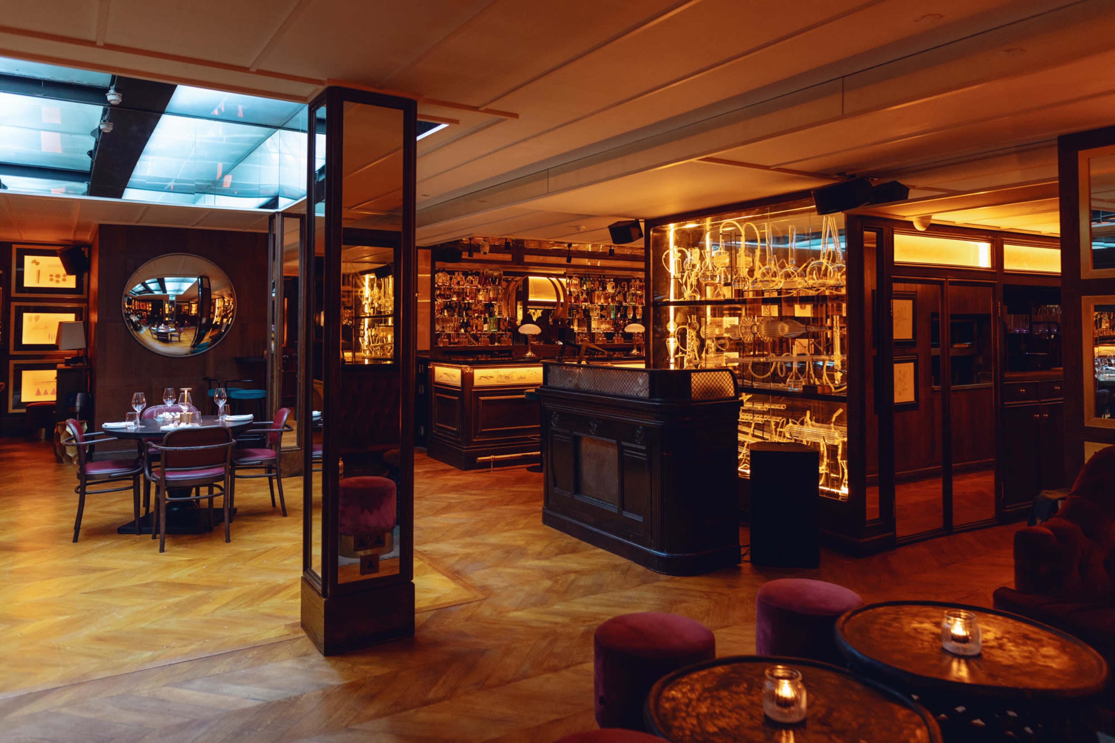 A stylish restaurant interior features a wooden bar, several tables, and shelves stocked with bottles of spirits under warm lighting.