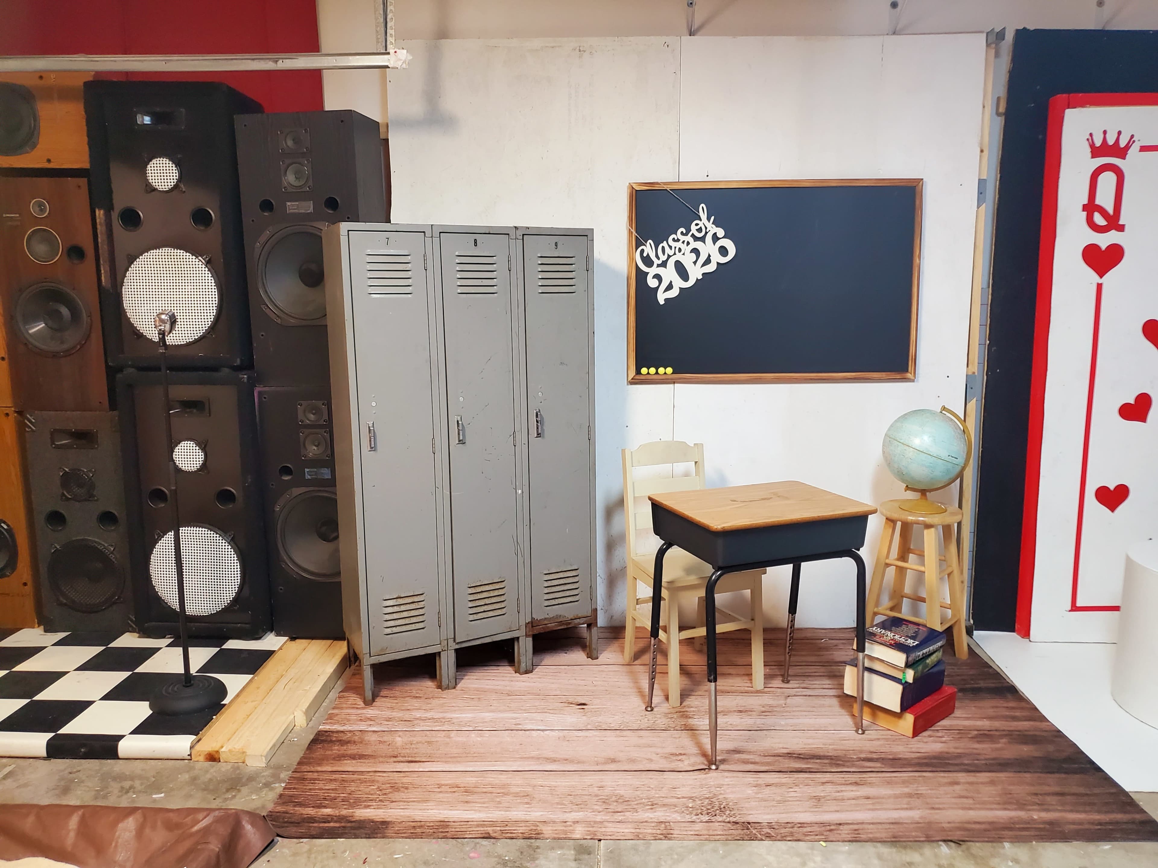 The image shows an indoor setup designed to resemble a school environment, featuring lockers, a chalkboard, a desk with a globe, and stacks of books beside a microphone and speakers.