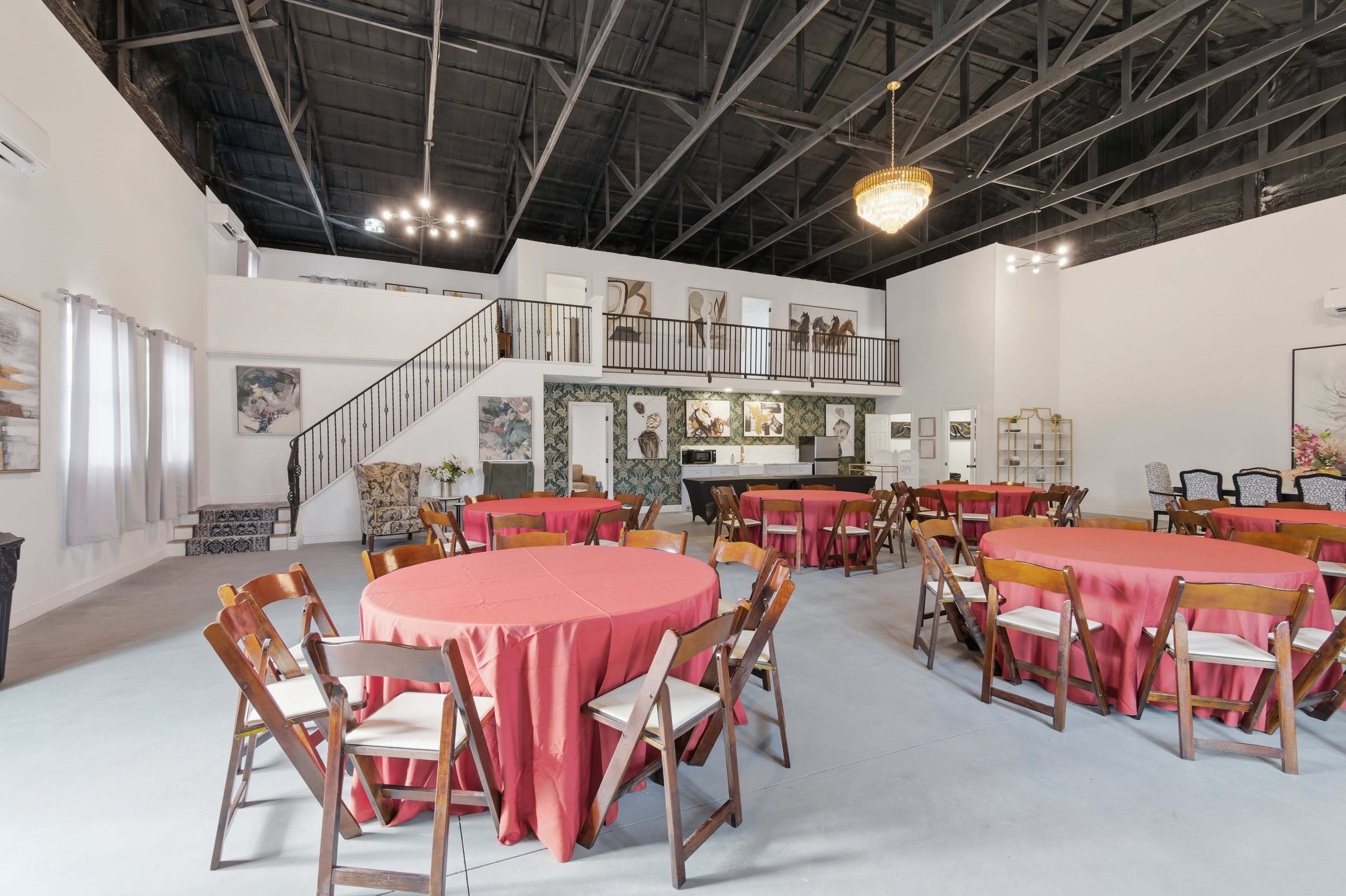 The image shows a spacious event room set with round tables covered in red tablecloths, surrounded by wooden chairs, and featuring a staircase and bar area in the background.