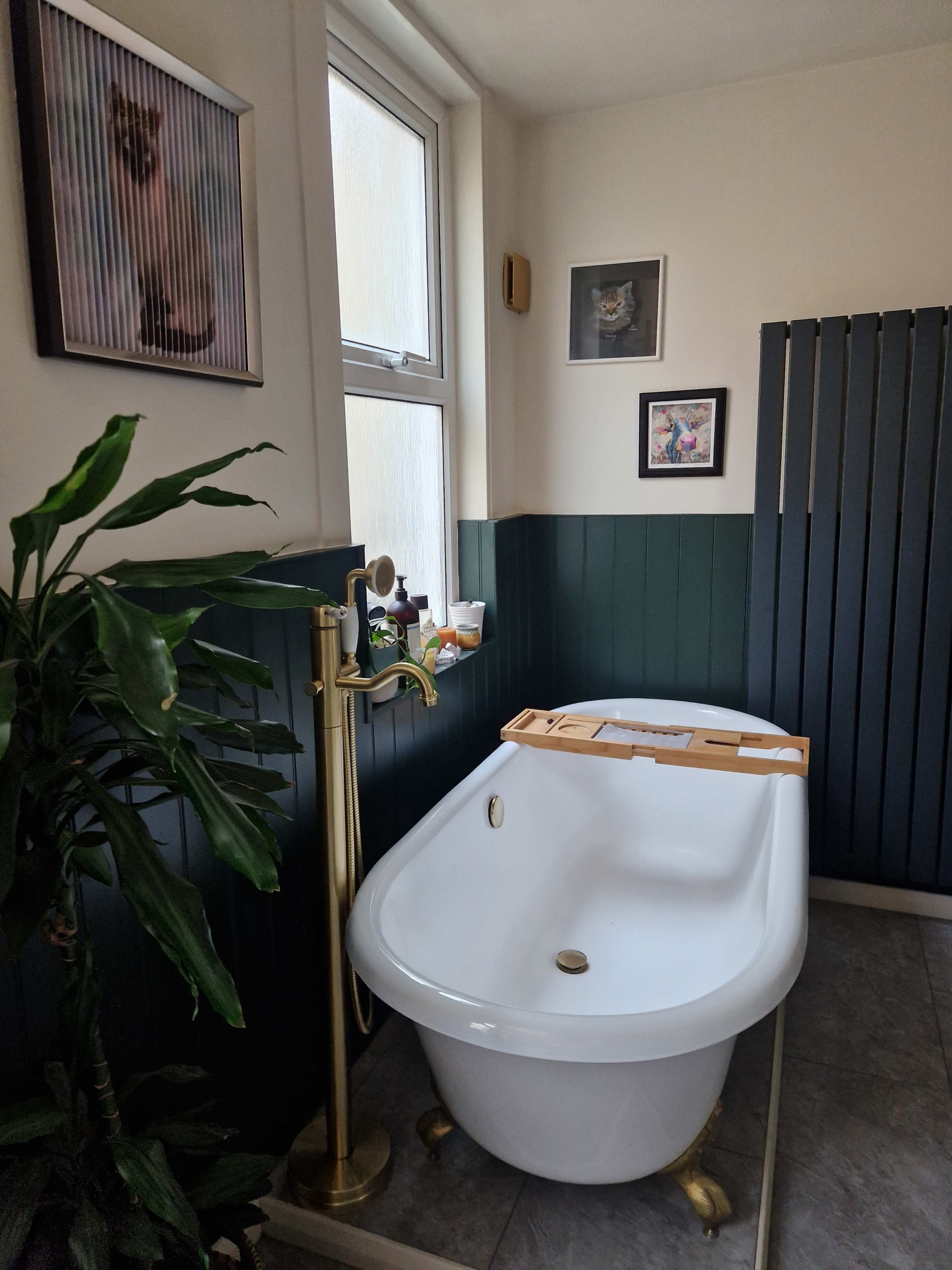 A white freestanding bathtub in a bathroom with green-paneled walls and framed artwork on the walls.