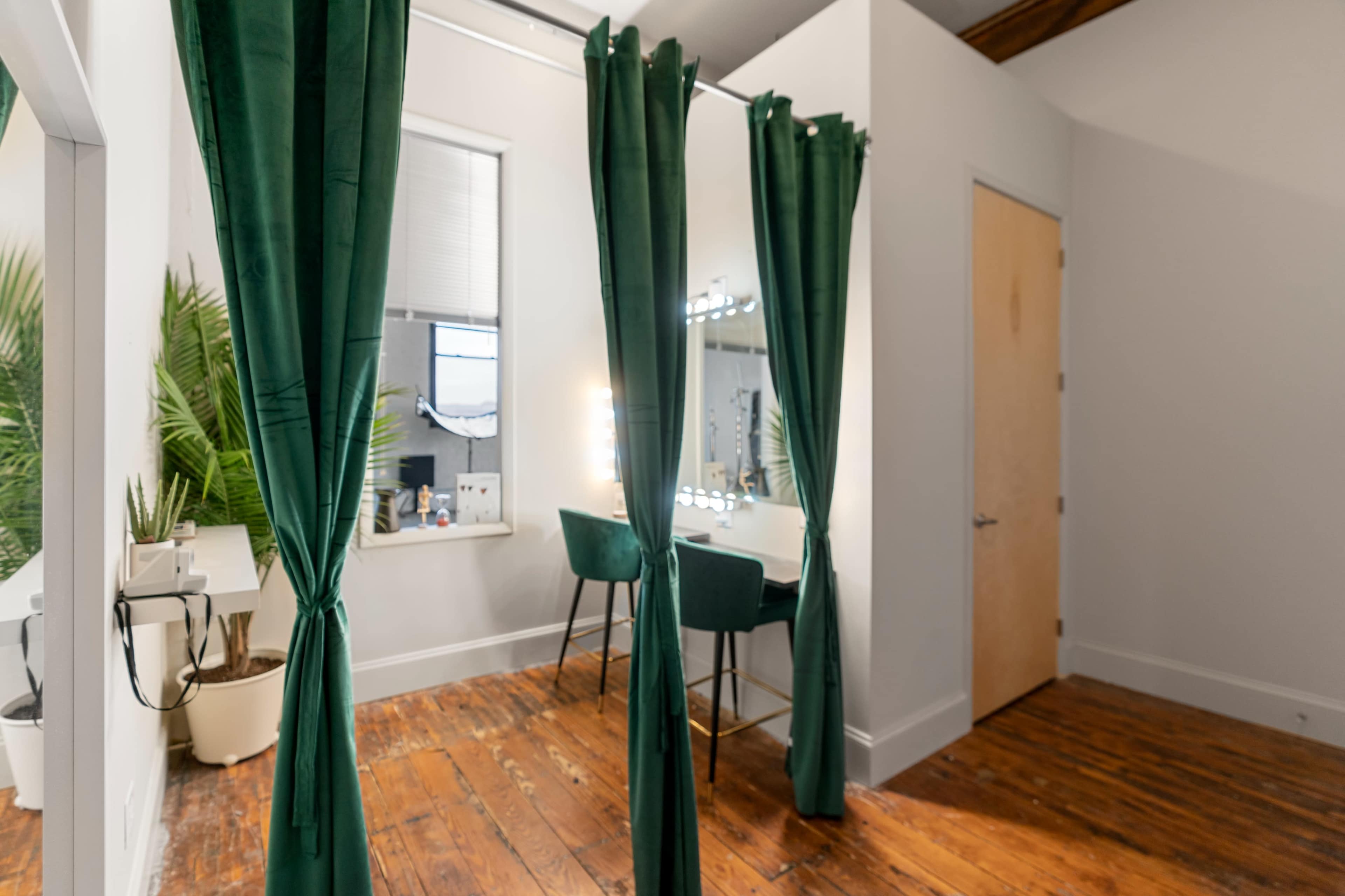 A spacious room features two green curtains dividing areas, with a vanity and chairs illuminated by mirrored lights in the background.