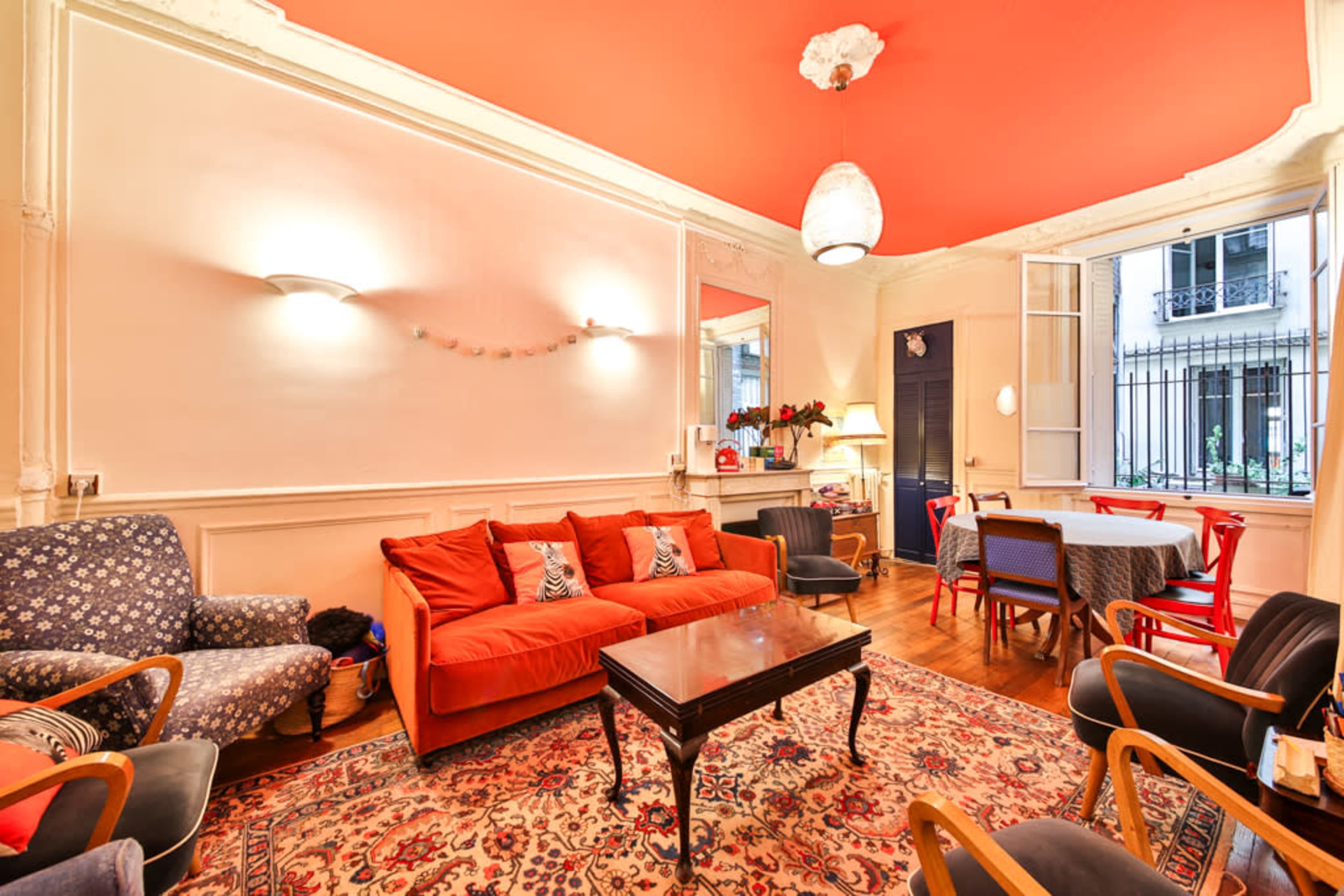 The image depicts a living room with a red ceiling, featuring an orange sofa, a patterned rug, and various seating options arranged around a wooden coffee table.