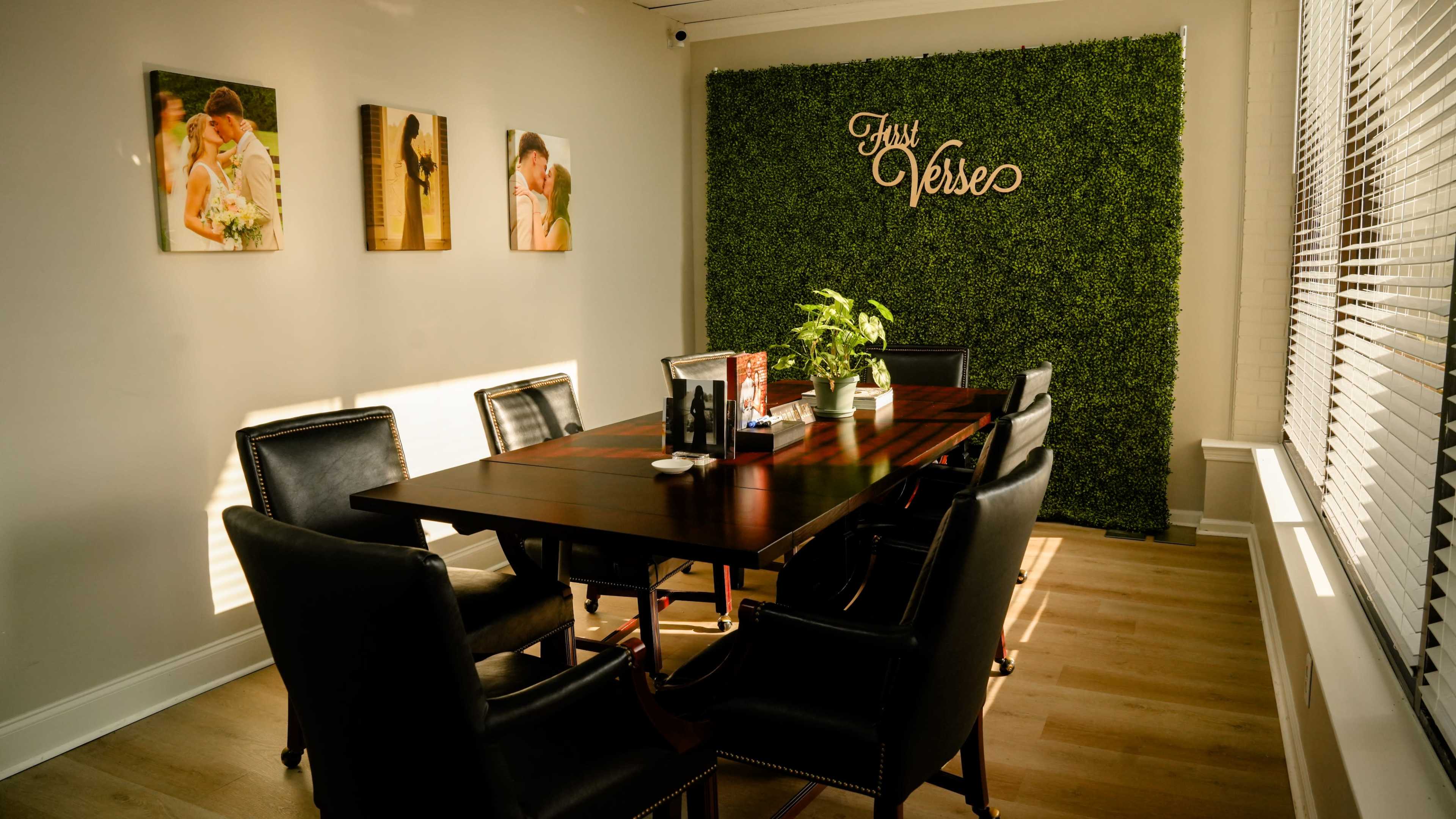 The image shows a conference room with a large wooden table surrounded by black leather chairs, featuring a green, plant-covered wall with the words "True Verse" displayed.