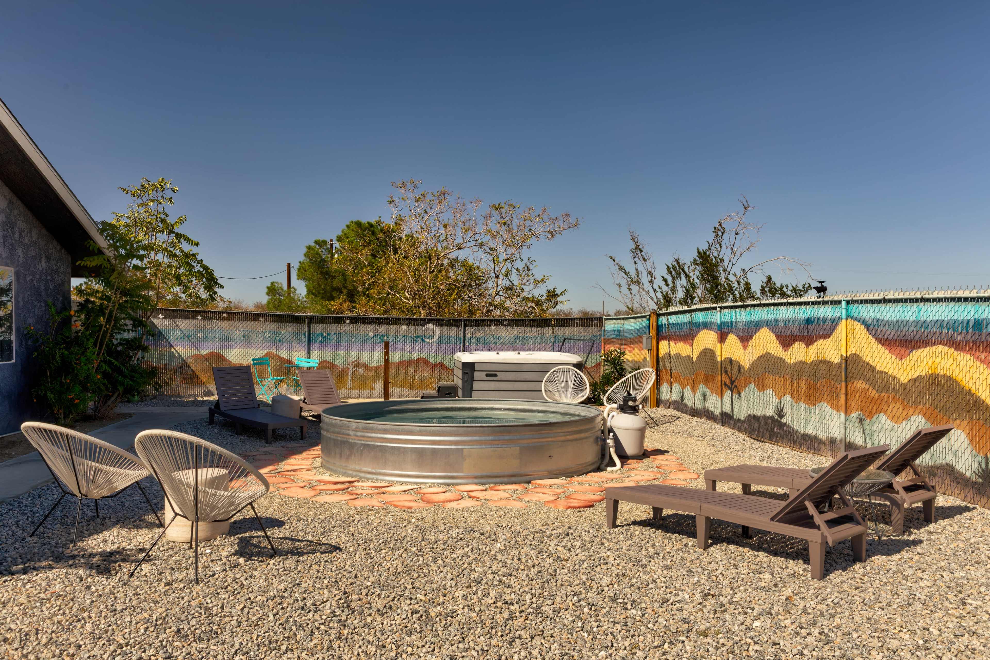The image shows a circular metal hot tub surrounded by gravel, with lounge chairs and a colorful muraled fence in the background.