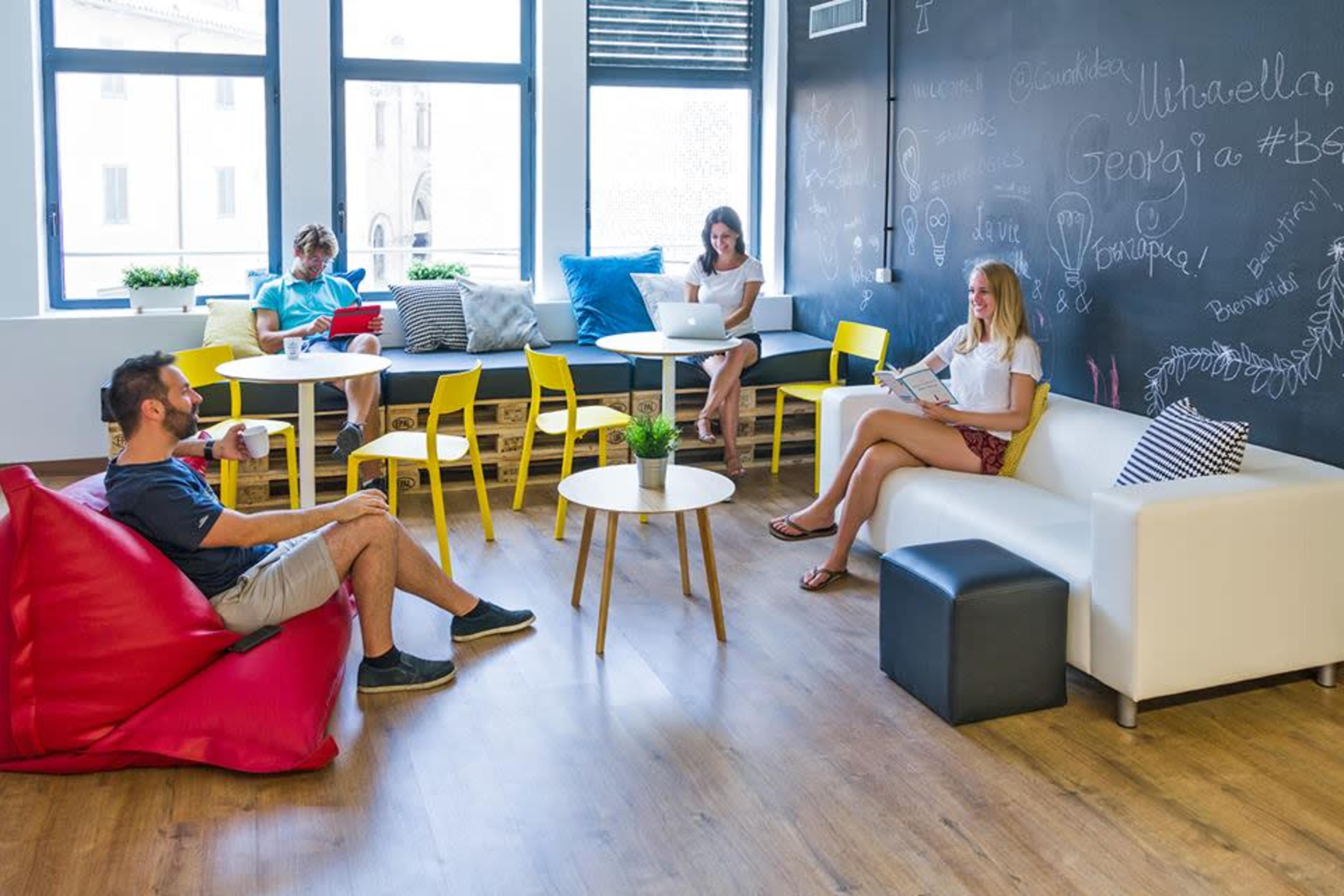 A group of four people is working and relaxing in a modern, brightly lit lounge area with colorful furniture and a chalkboard wall.