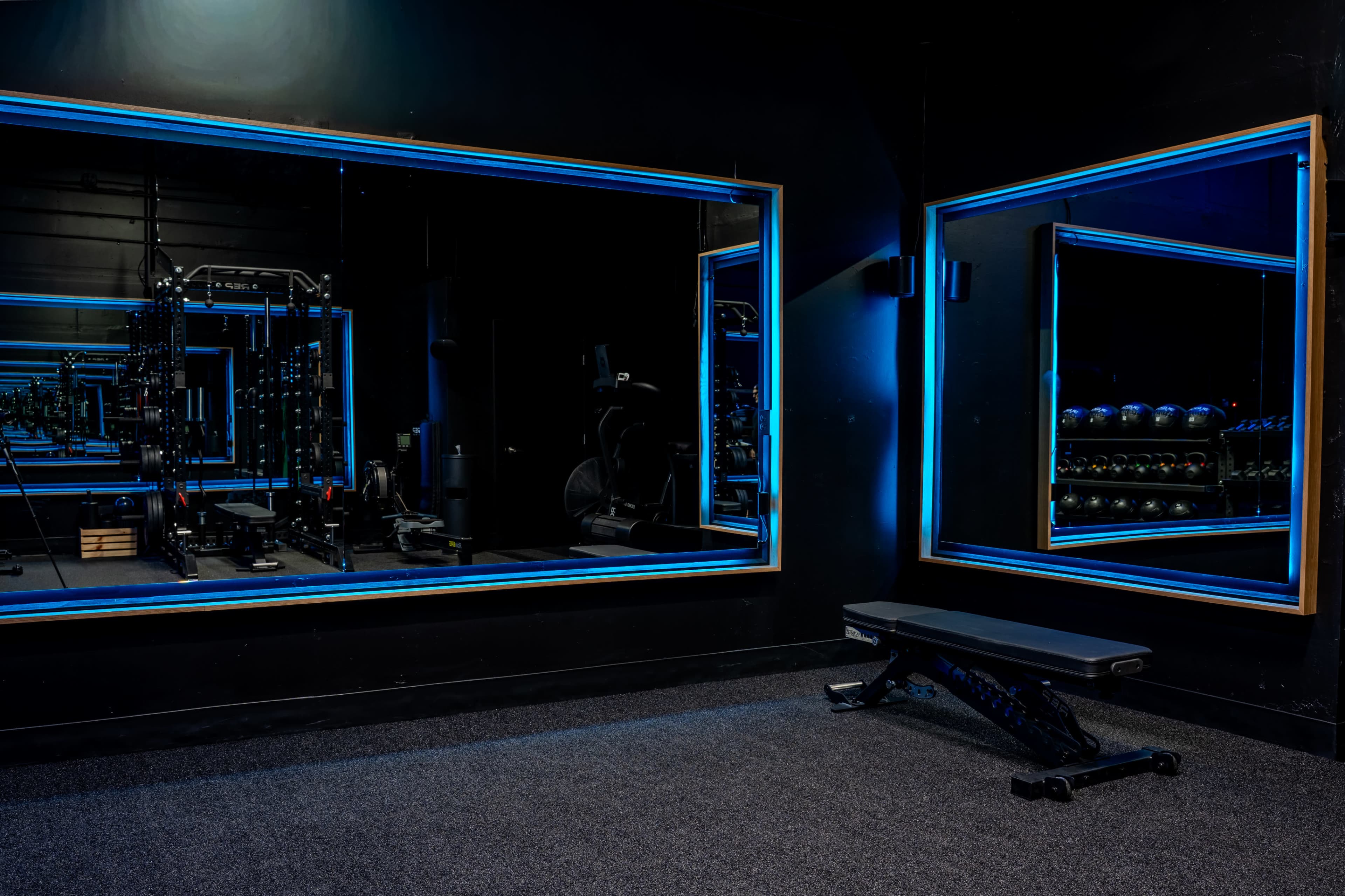 The image shows a gym interior with two illuminated mirrors, a weight bench, and various exercise equipment in a dark setting.