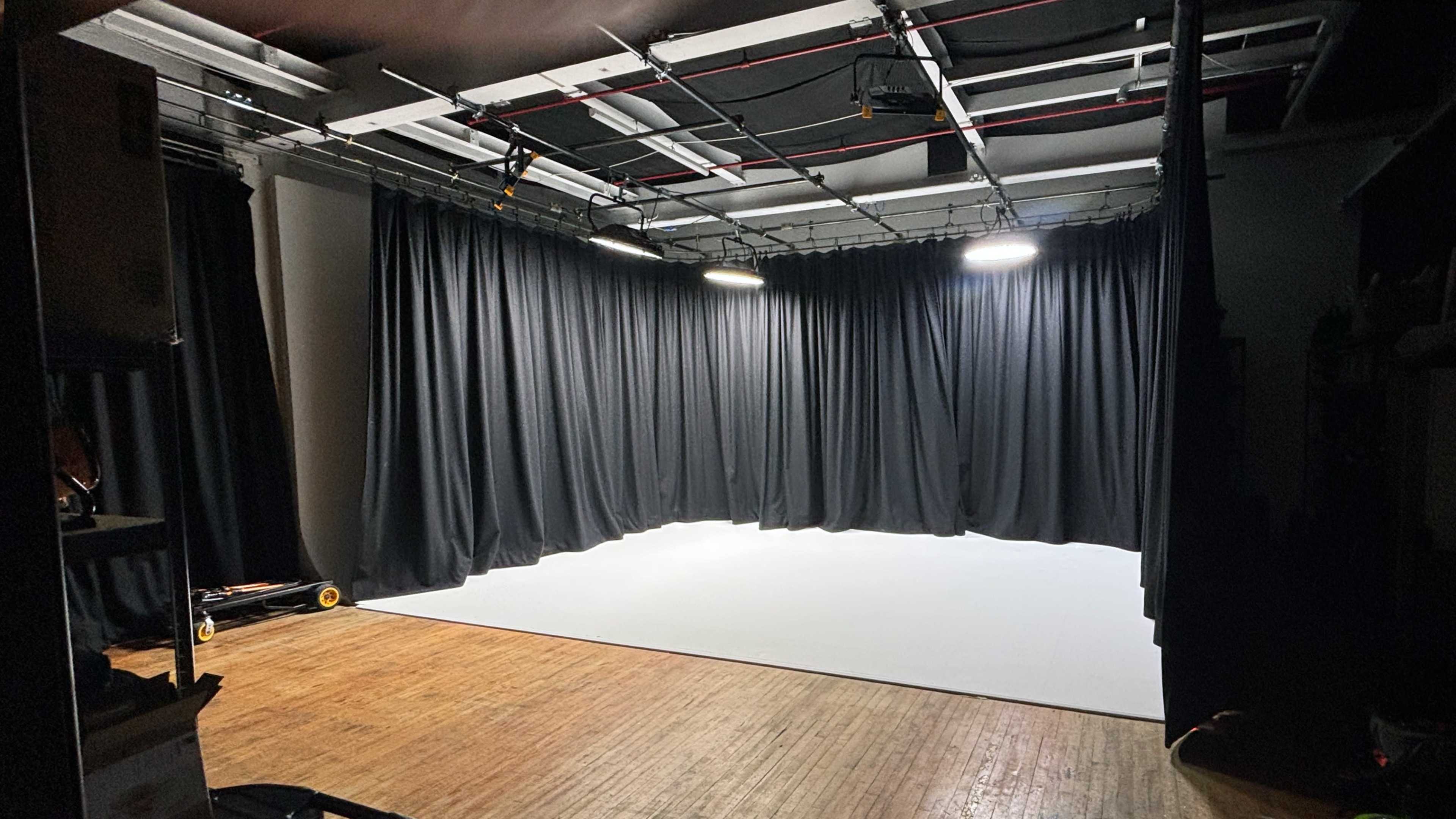 The image shows a blank stage area with black curtains draped along the walls and overhead lights illuminating a white floor.