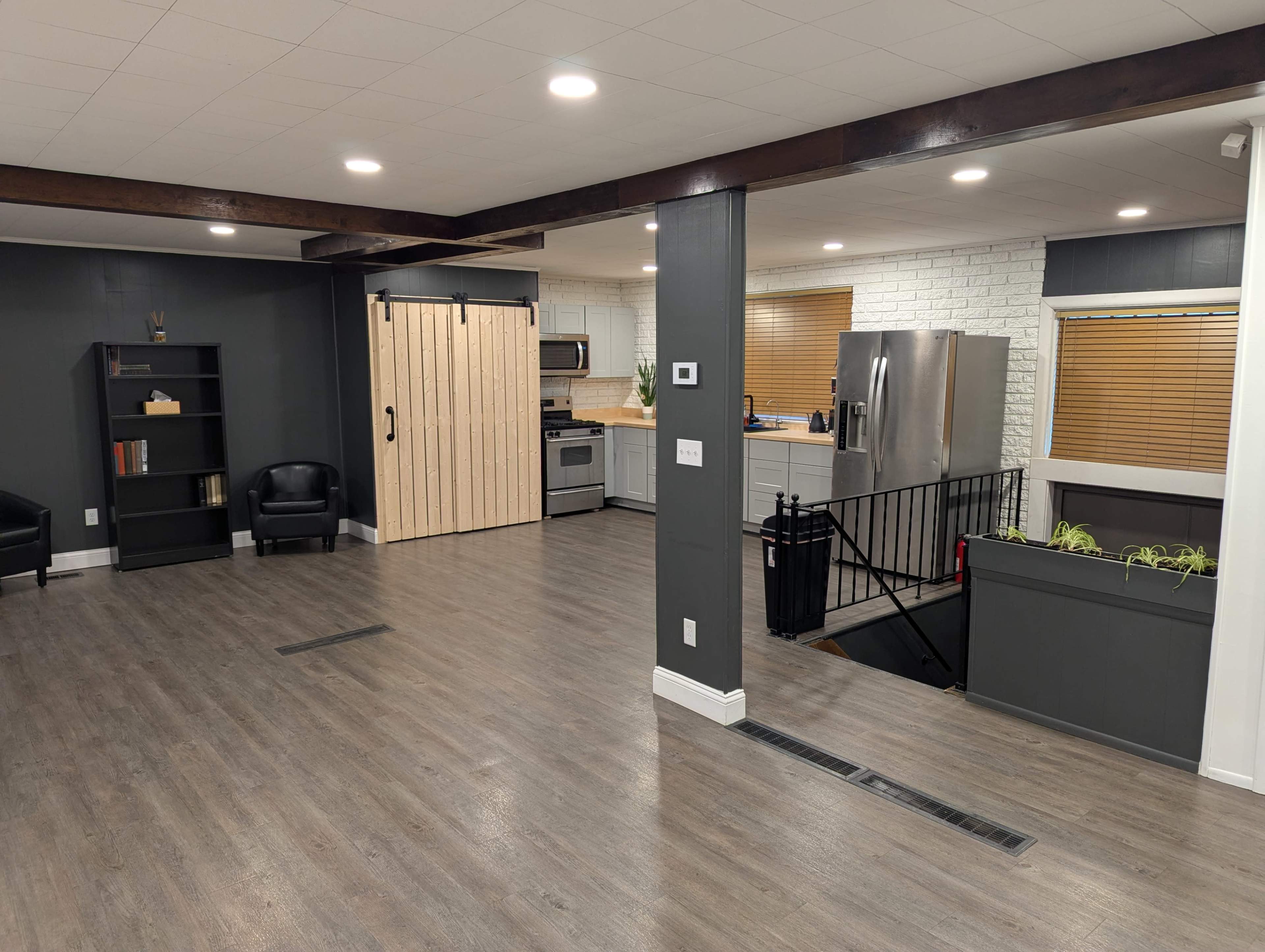 The image shows a spacious, modern living area with wooden accents, a kitchen in the background, and a staircase leading to a lower level.