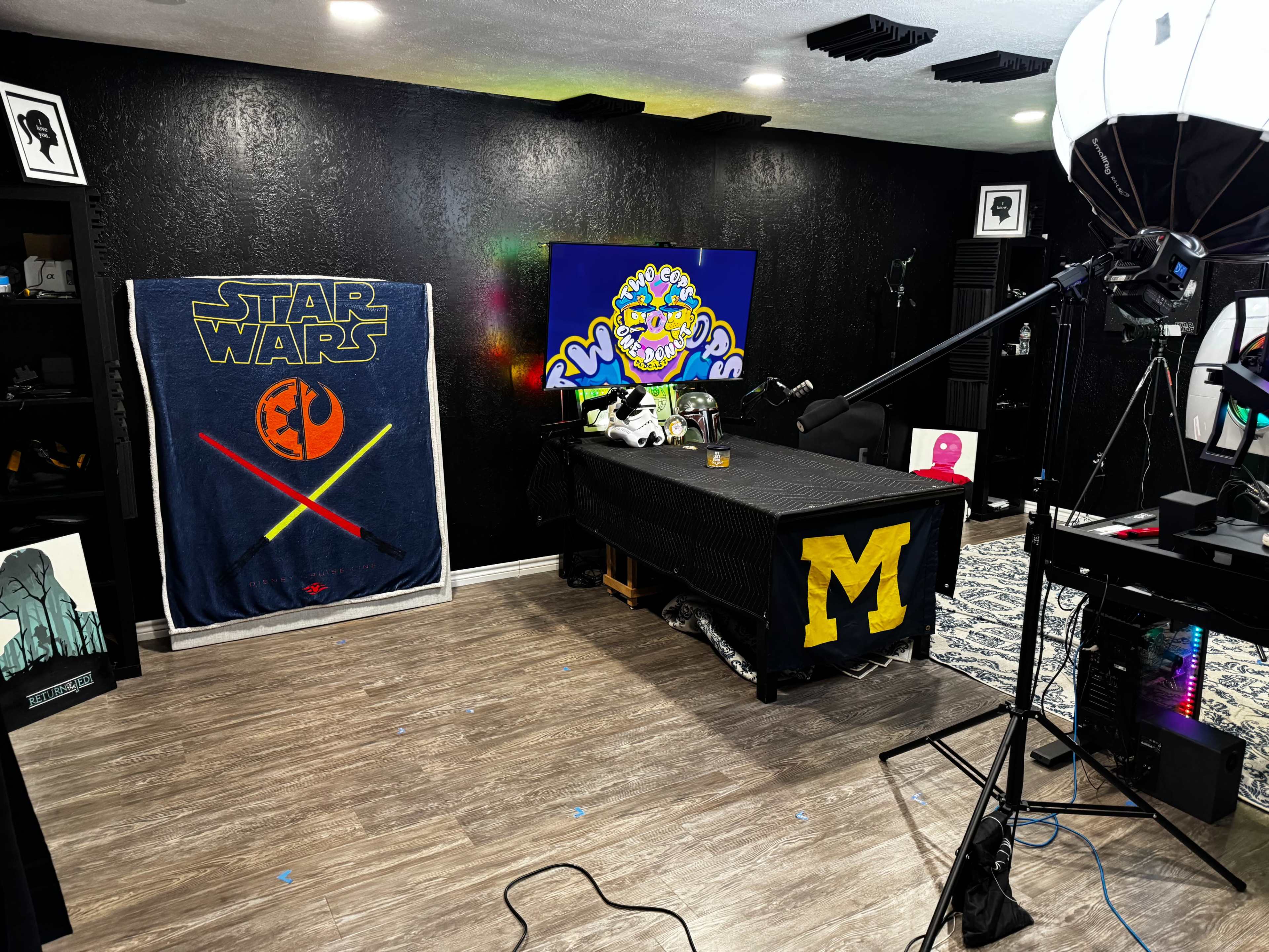 The image shows a room with a black wall, a Star Wars blanket draped on a wall, and a table adorned with a Michigan logo, along with various items and lighting equipment scattered around.