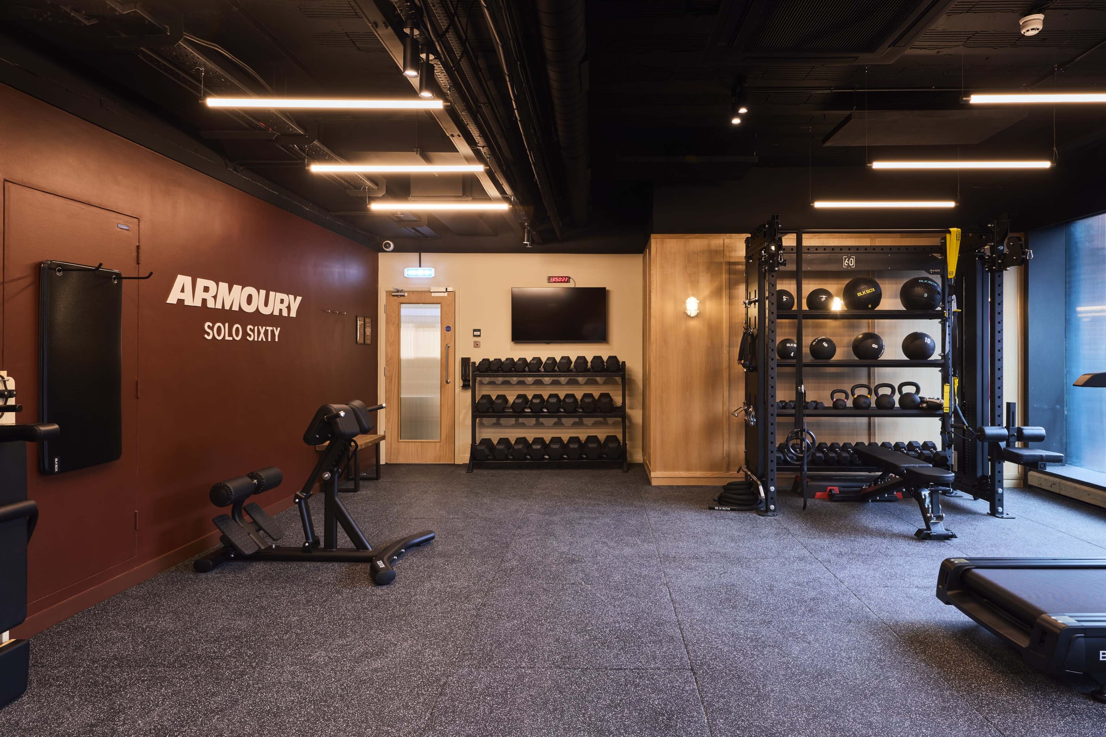 The image shows a modern gym interior with equipped workout areas, including weight storage, exercise machines, and a wall adorned with the word "ARMOURY."