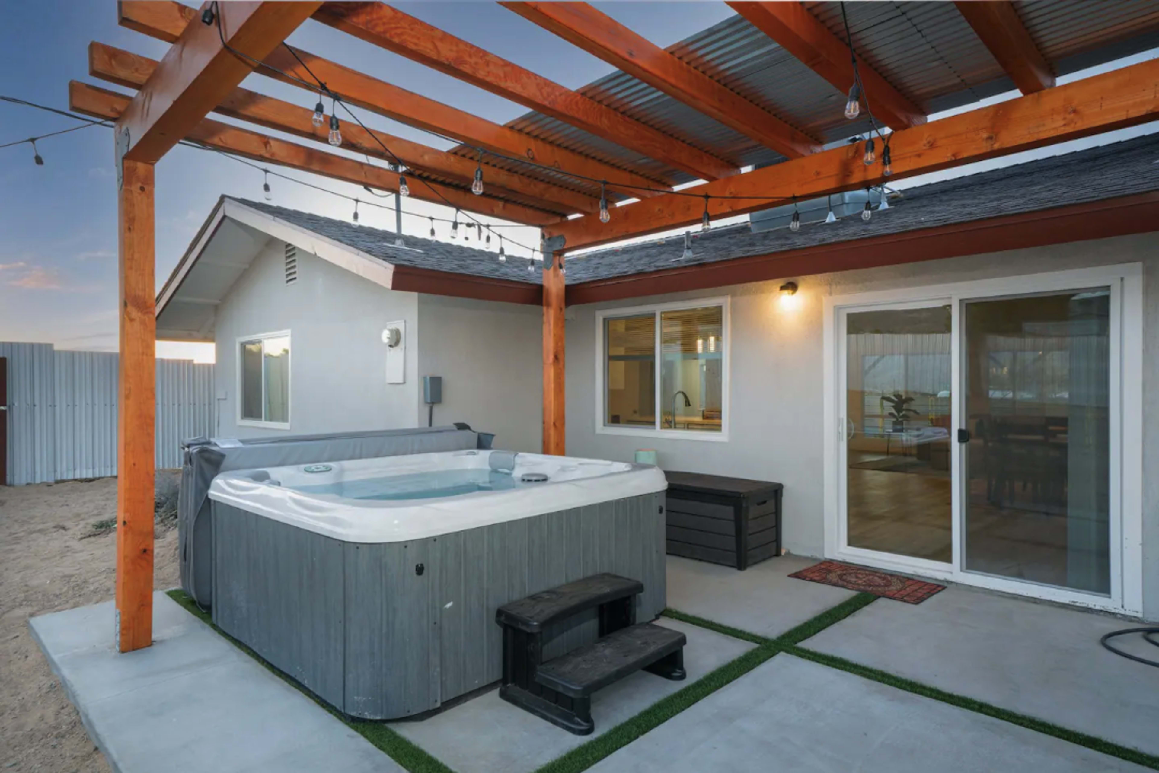 A hot tub situated under a wooden pergola beside a modern house with sliding glass doors and outdoor string lights.