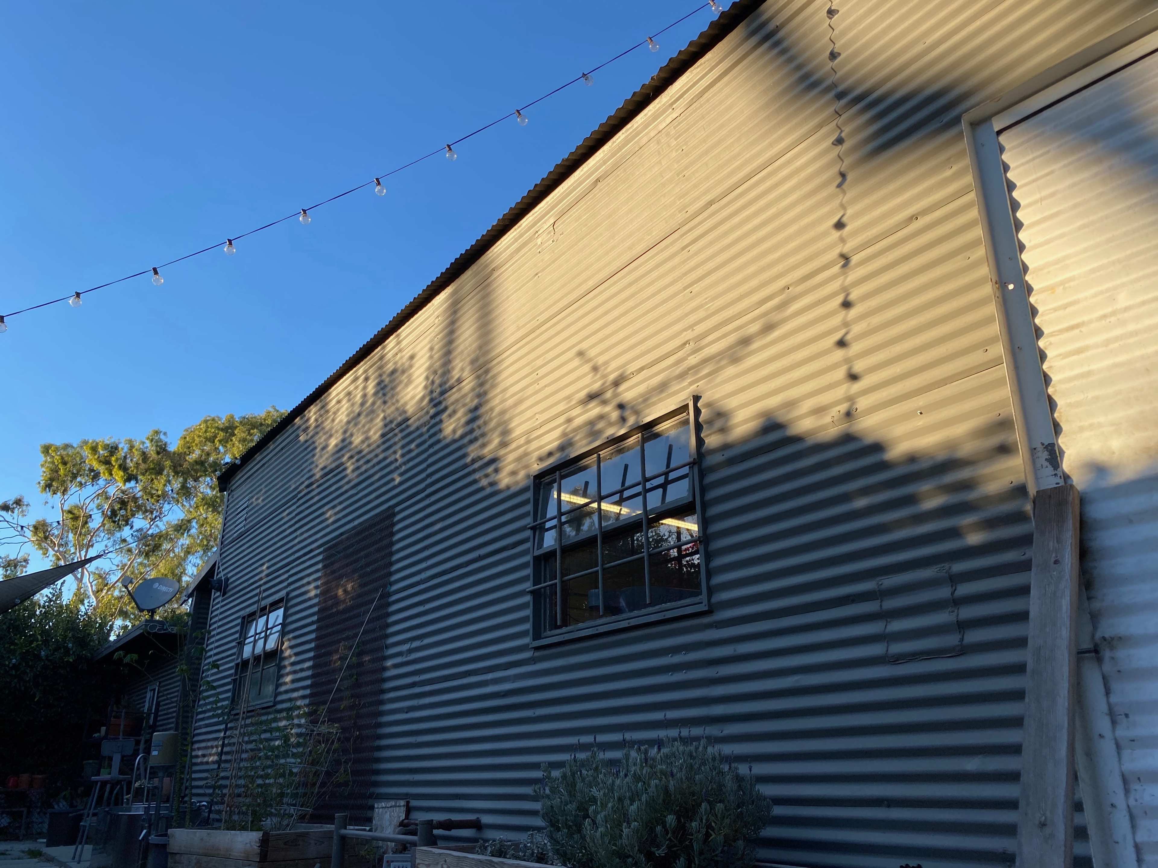 The image shows a corrugated metal building with a large window and shadows cast on its side, under a clear blue sky.