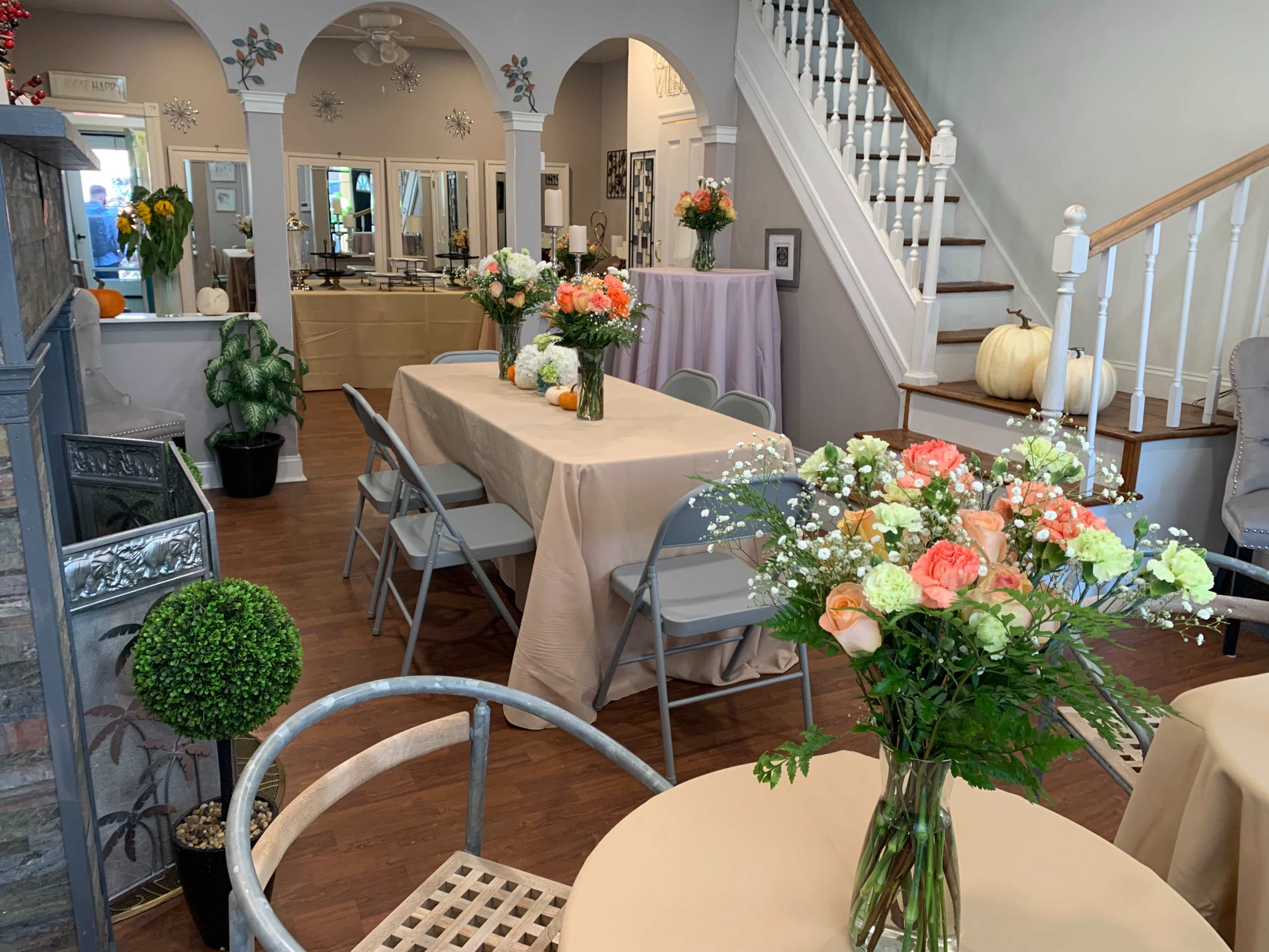 A brightly lit event space featuring several tables dressed with cream tablecloths and floral centerpieces, a staircase in the background, and decorative plants throughout the room.