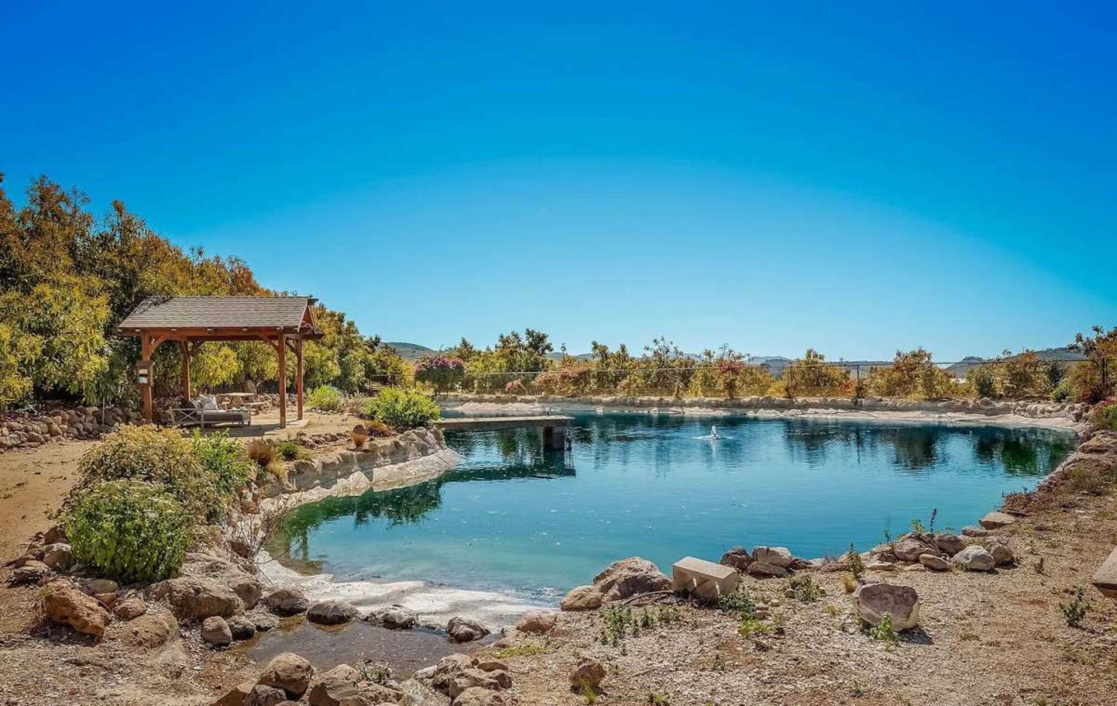 The image shows a clear pond surrounded by rocks and greenery, with a wooden gazebo nearby under a blue sky.