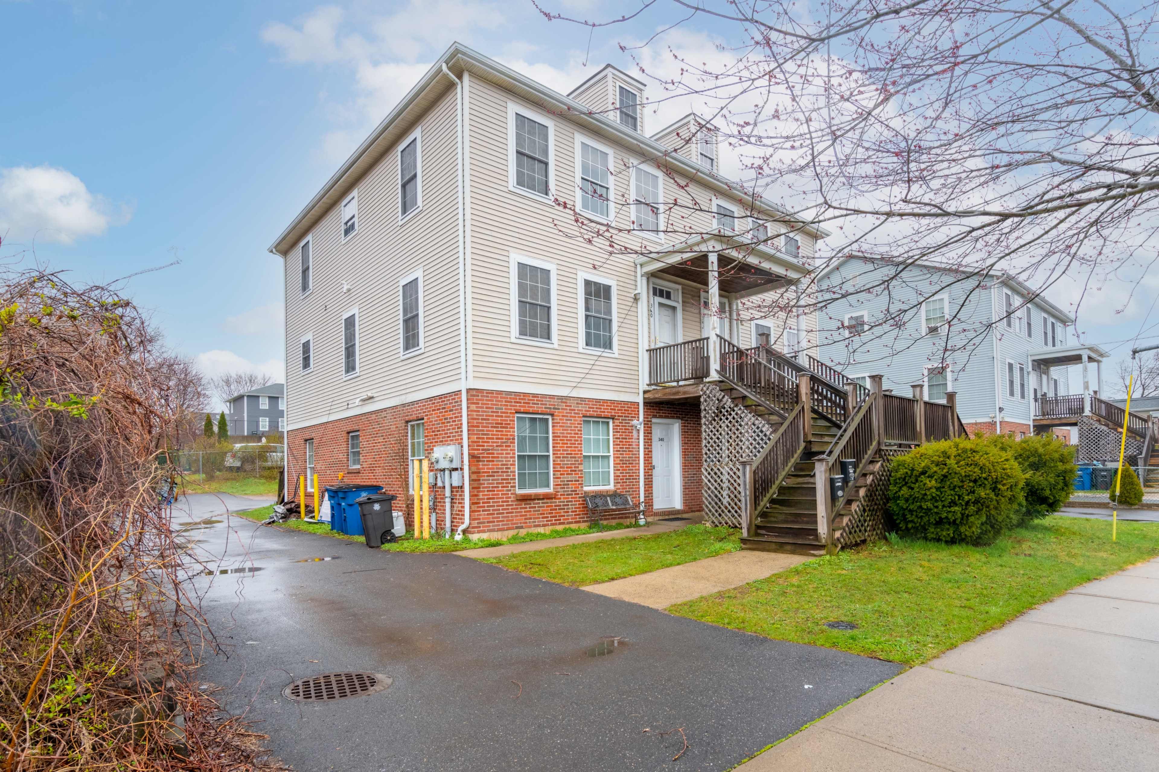 The image shows a three-story residential building with a brick and siding exterior, a staircase leading to a second-floor entrance, and parked refuse bins along a paved pathway.