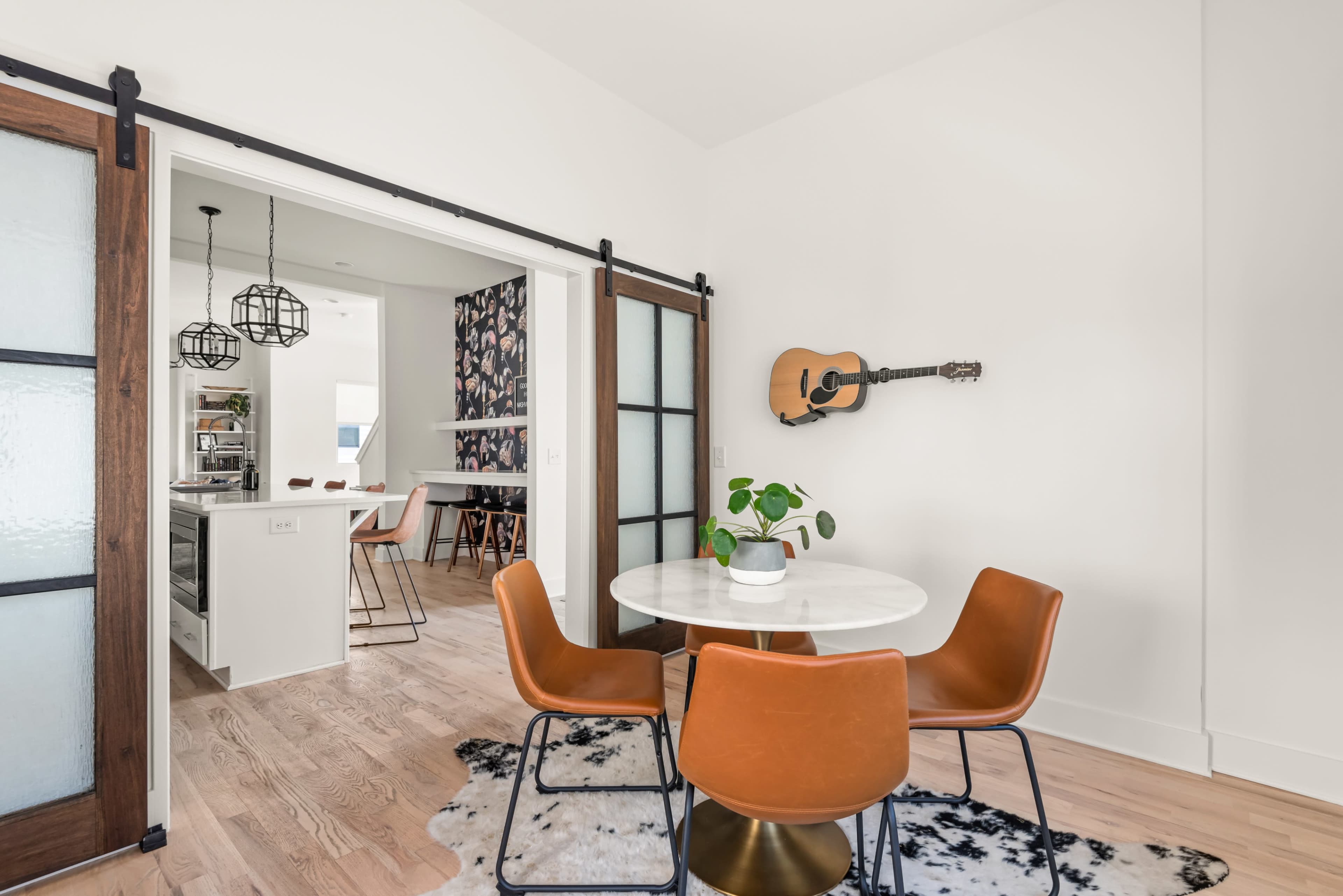 A round marble table with three orange chairs sits in a brightly lit dining area, adjacent to a kitchen visible through a sliding glass door.