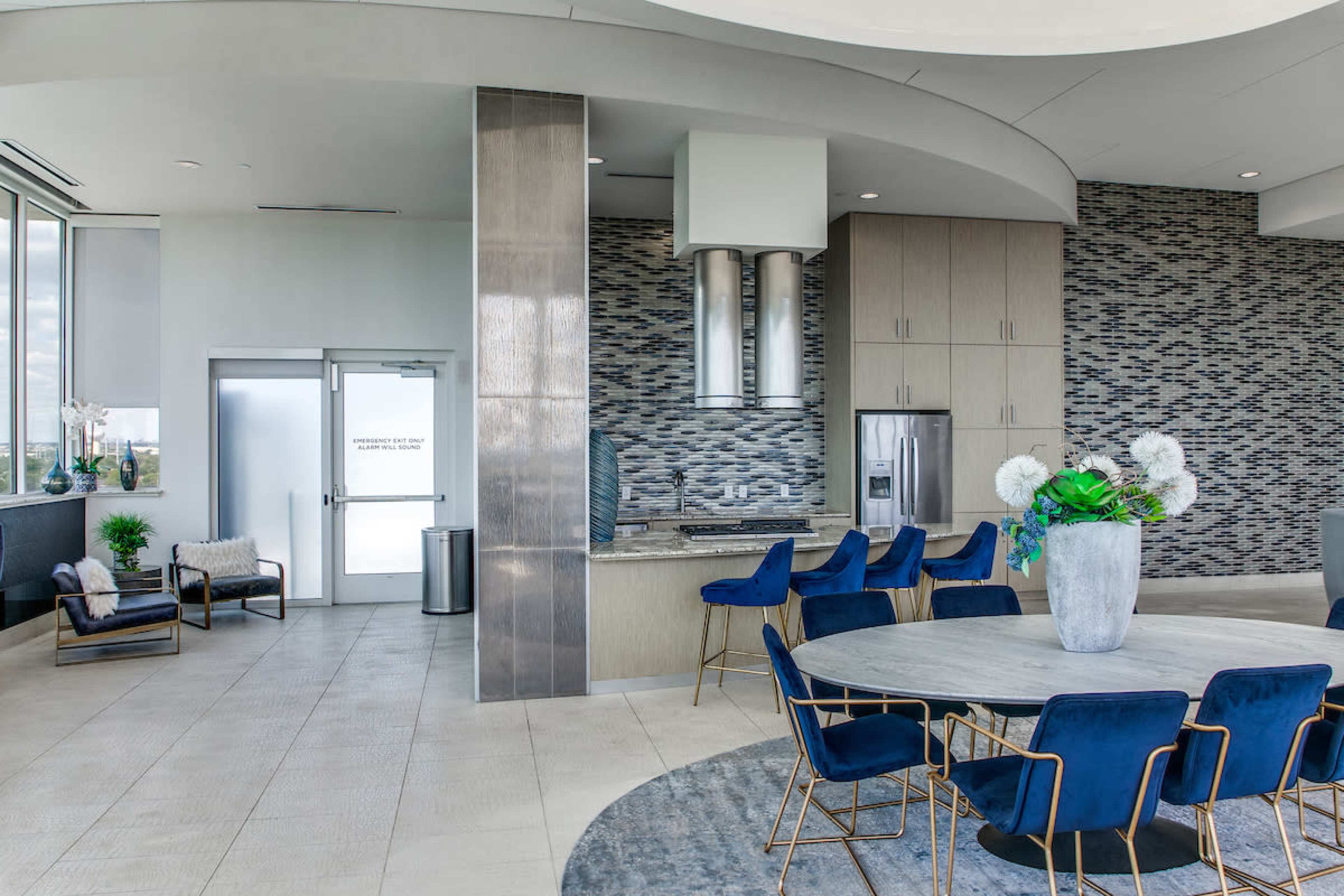 A modern kitchen and dining area features a round table with blue chairs, a large vase of flowers, and stainless steel appliances against a patterned tile backsplash.