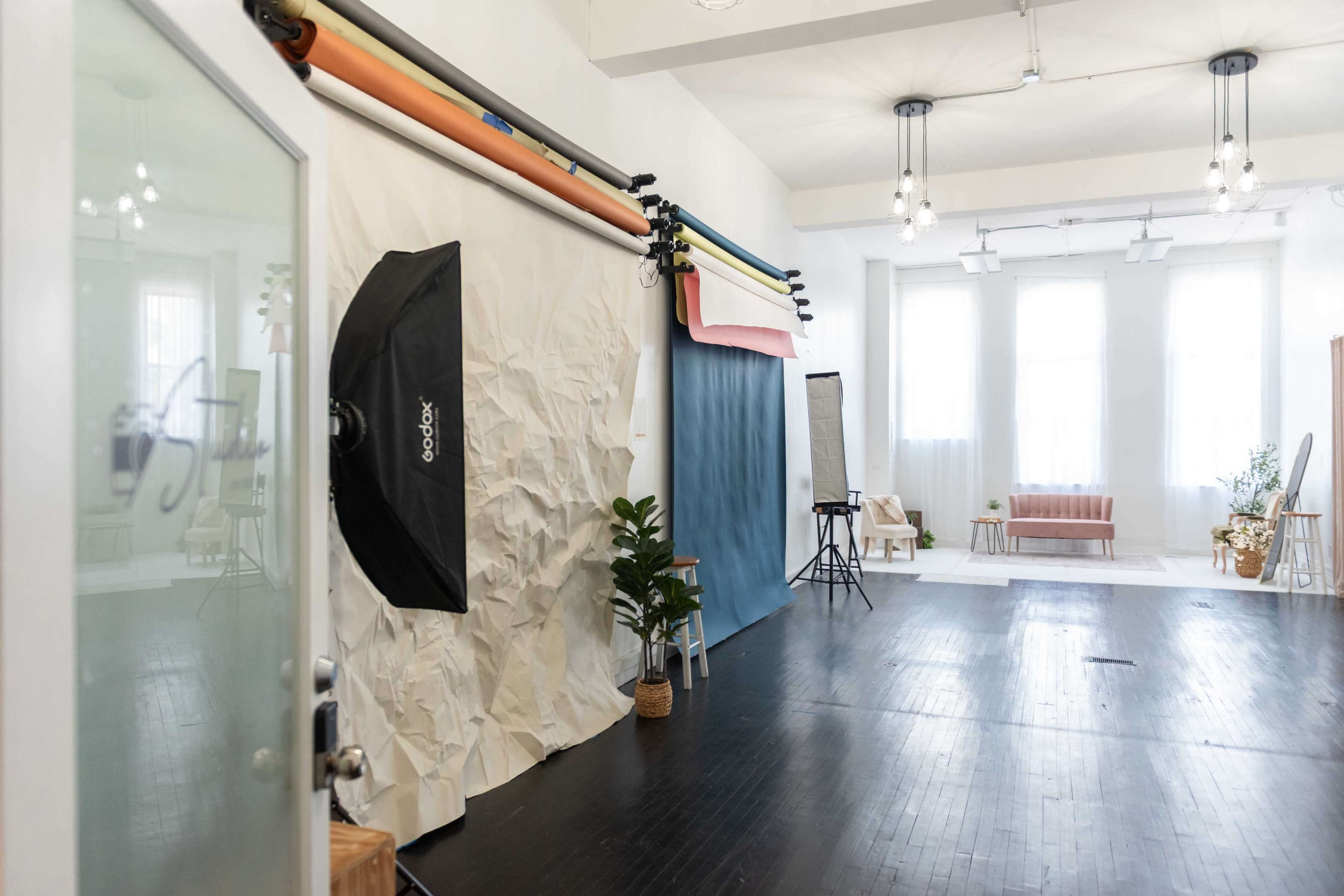The image shows a spacious photography studio featuring a backdrop setup with colorful paper rolls, a softbox light, and a seating area with a couch.