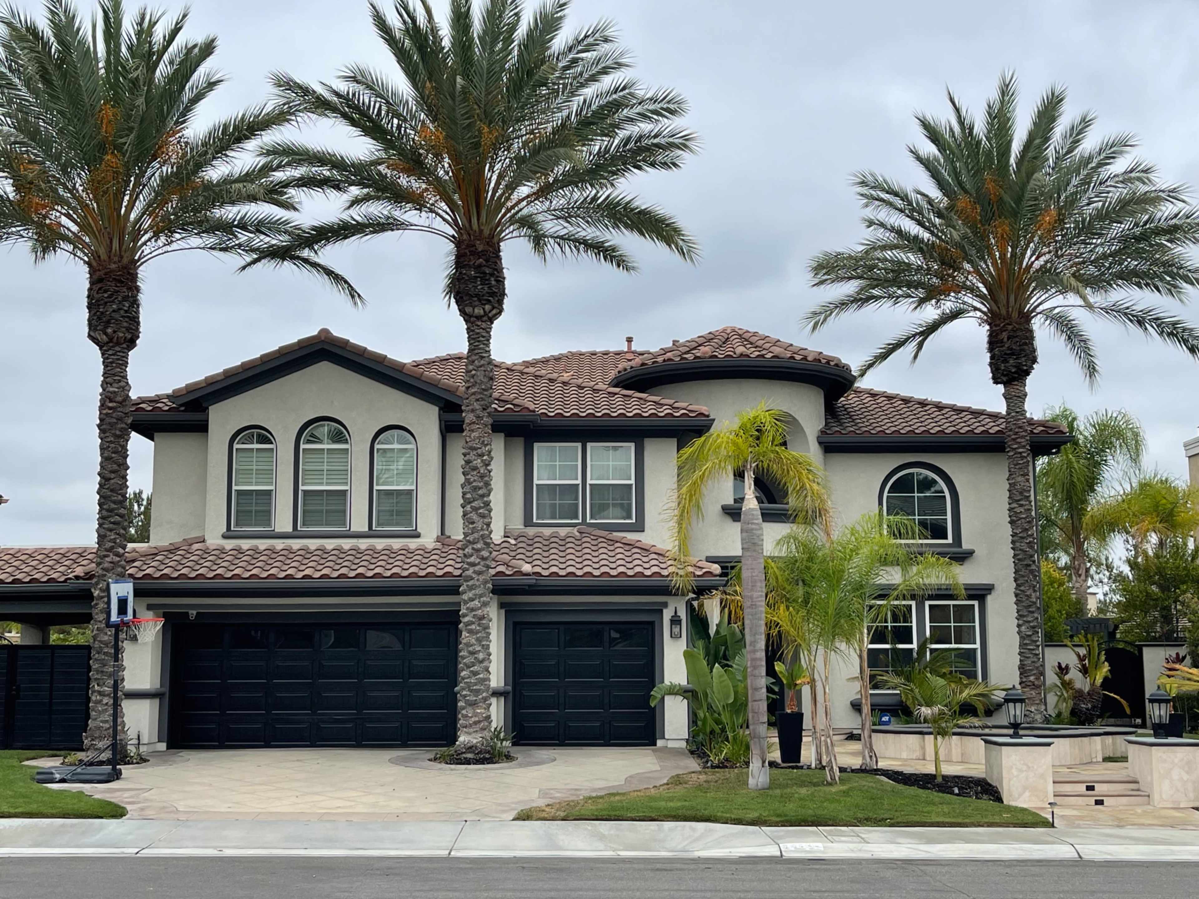 A large two-story house with a tiled roof, flanked by palm trees, features a three-car garage and a landscaped front yard.