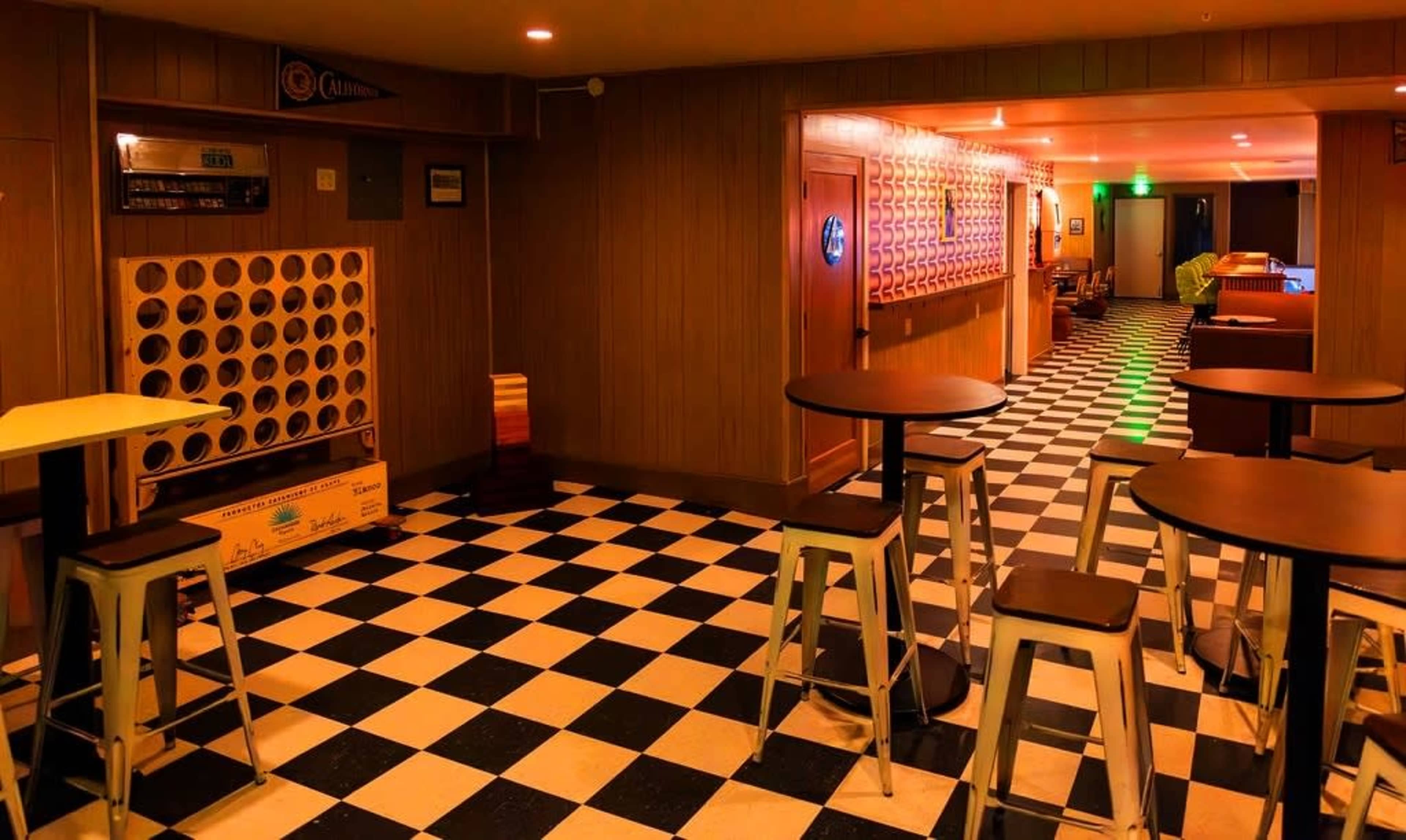 The image shows a hallway with a checkered black-and-white floor, featuring tables and chairs on one side and ambient lighting along the walls.