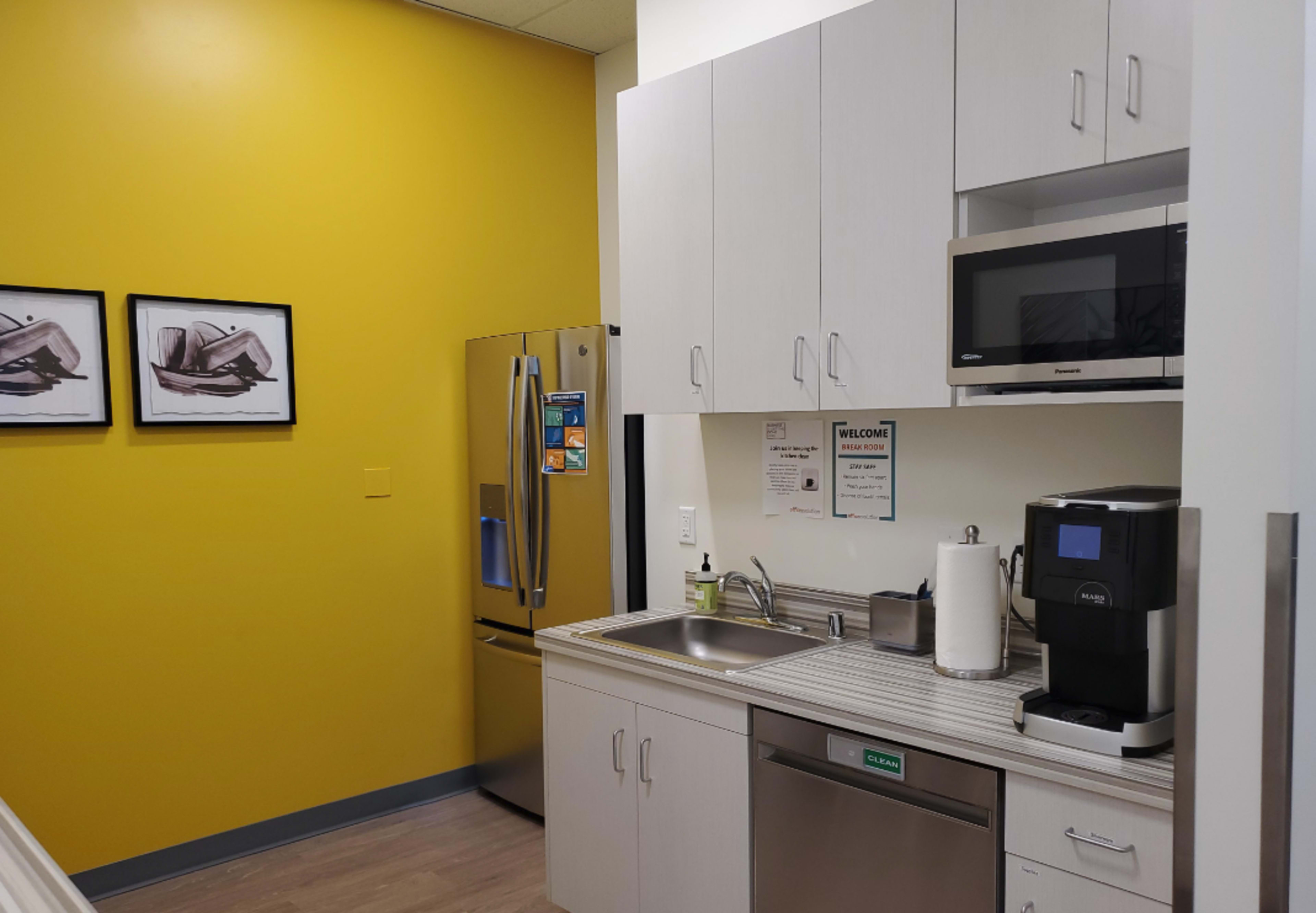 The image shows a modern kitchen area with a yellow accent wall, stainless steel refrigerator, gray cabinets, a sink, and a coffee machine.