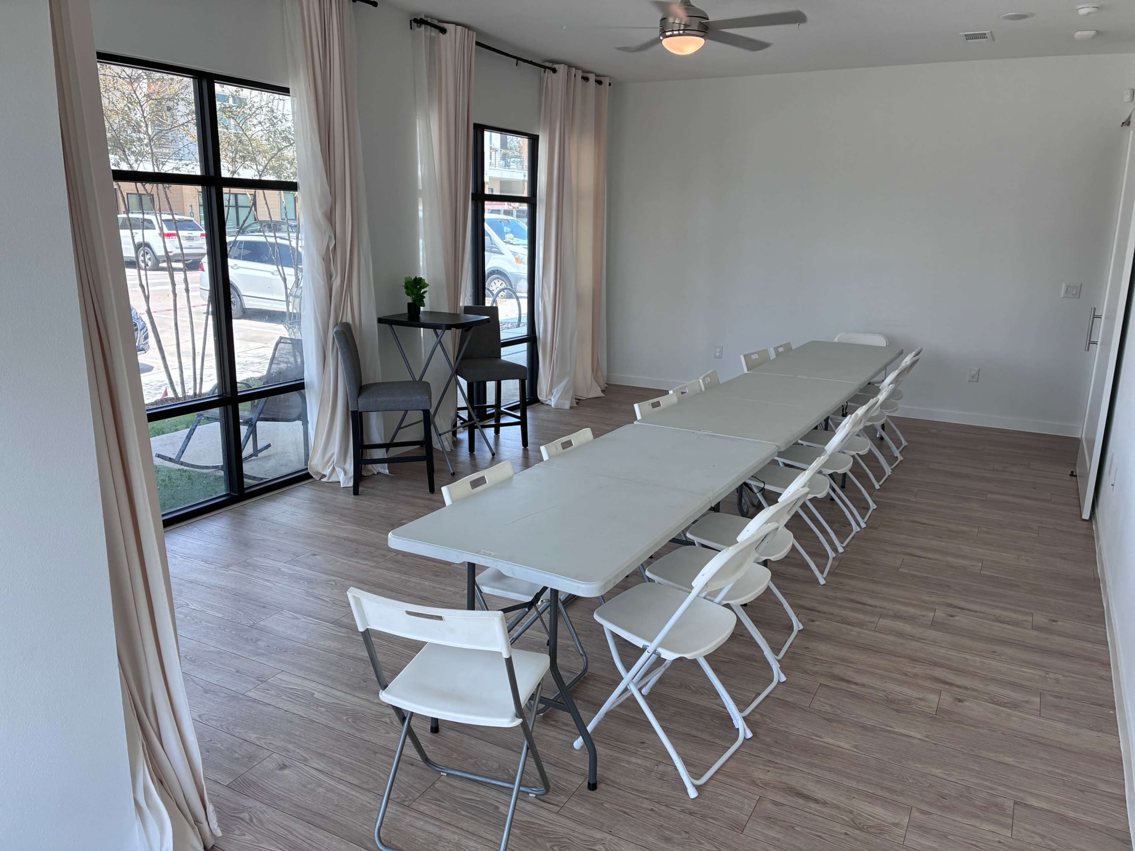 The image shows a well-lit room with a long table set up with folding chairs, large windows, and a small standing table in one corner.
