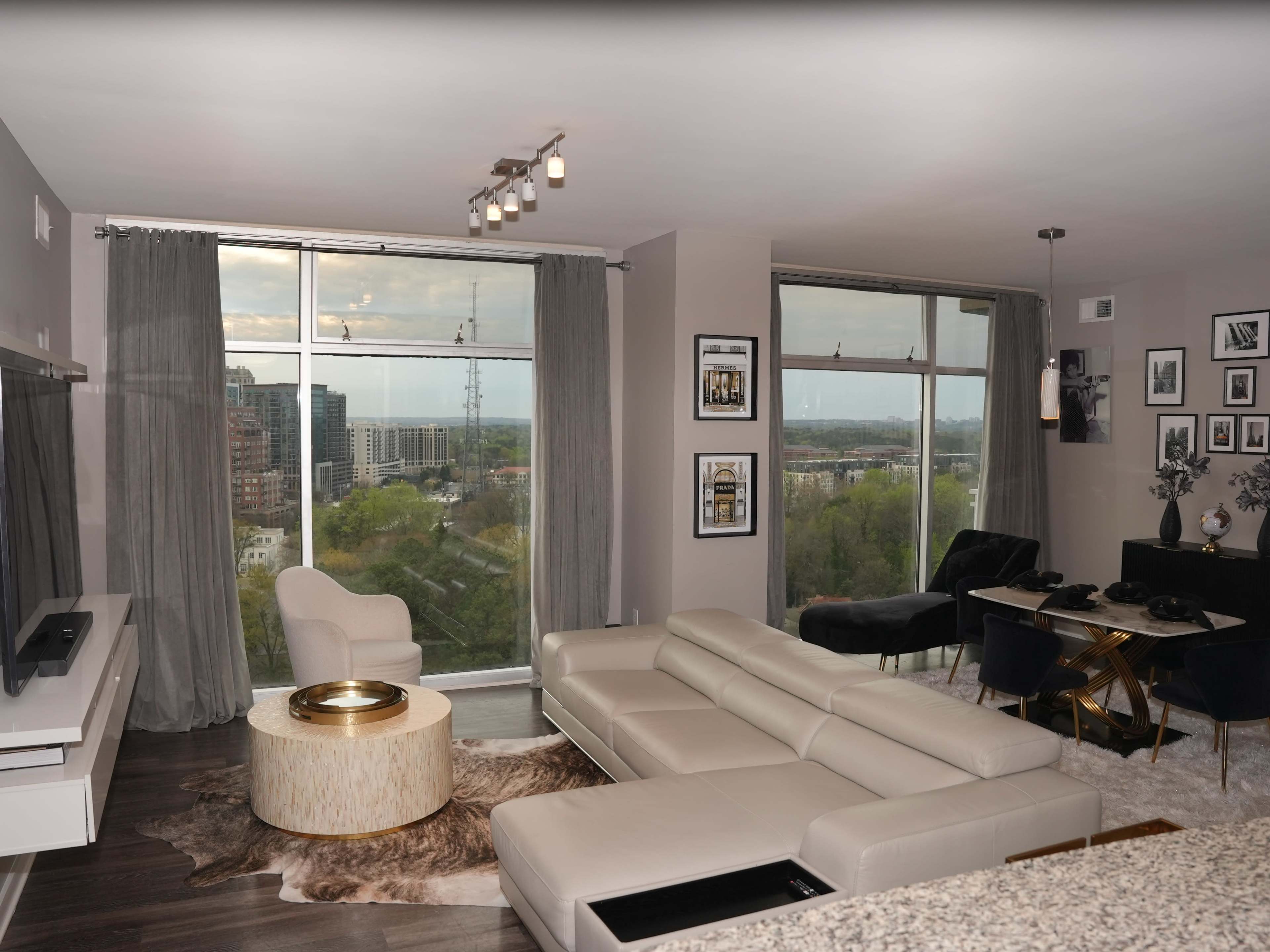 A modern living room features a large sectional sofa, a round coffee table, and a dining area, all with floor-to-ceiling windows showcasing a city view.