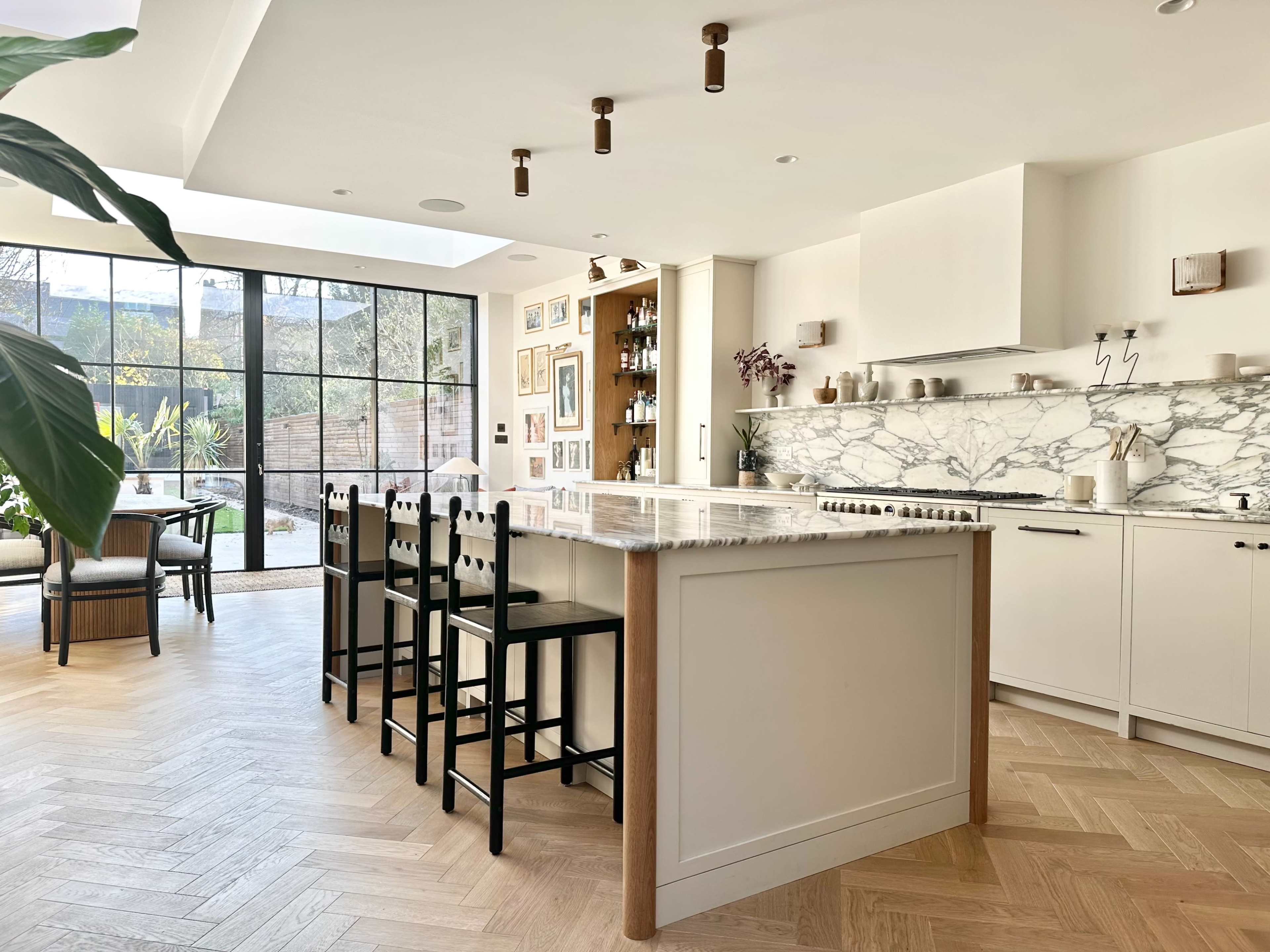 The image shows a modern kitchen with a marble countertop, black barstools, and large windows overlooking a garden.