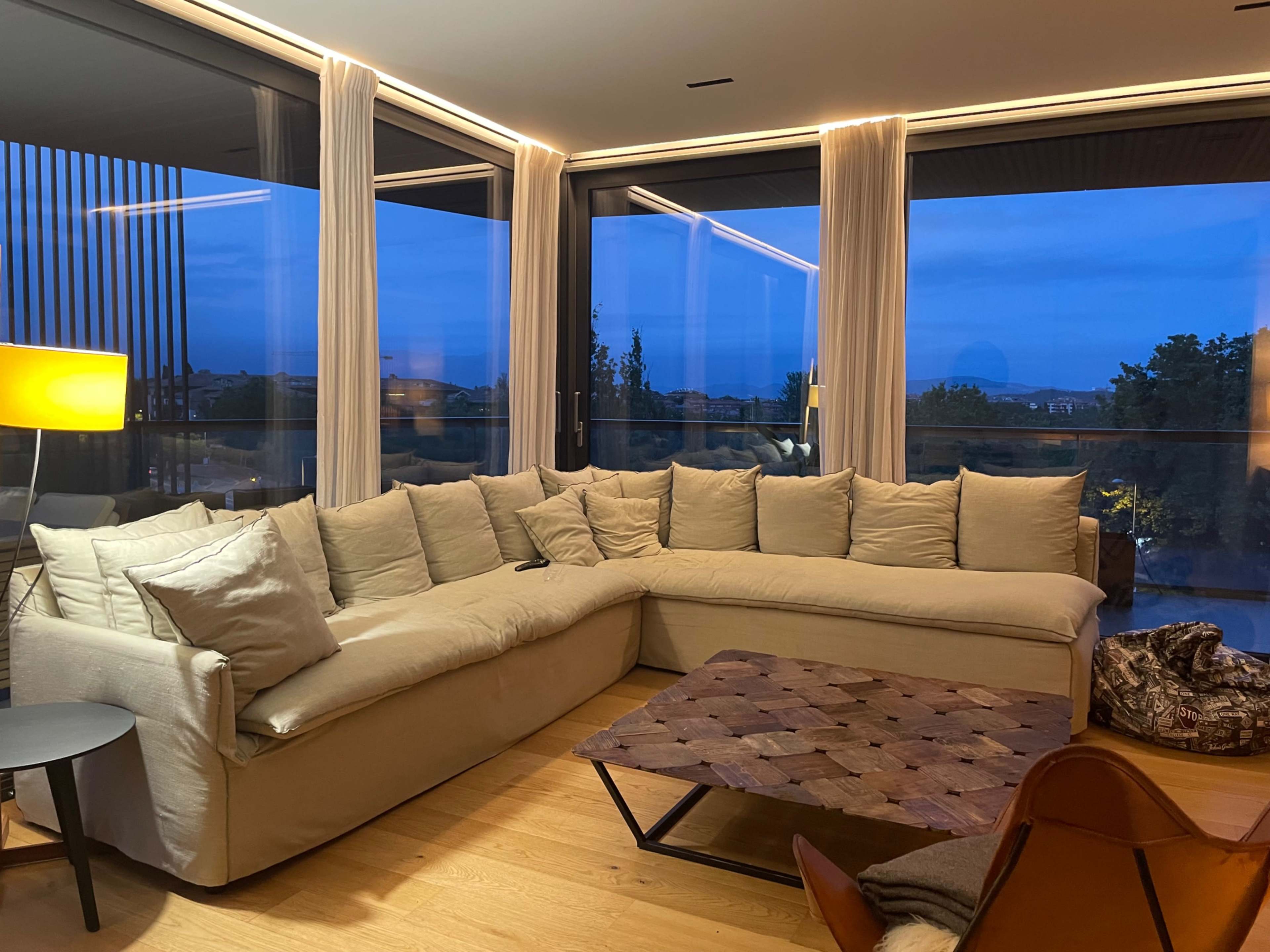 A spacious living room features a large sectional sofa, a wooden coffee table, a floor lamp, and large windows with a view of the evening sky.