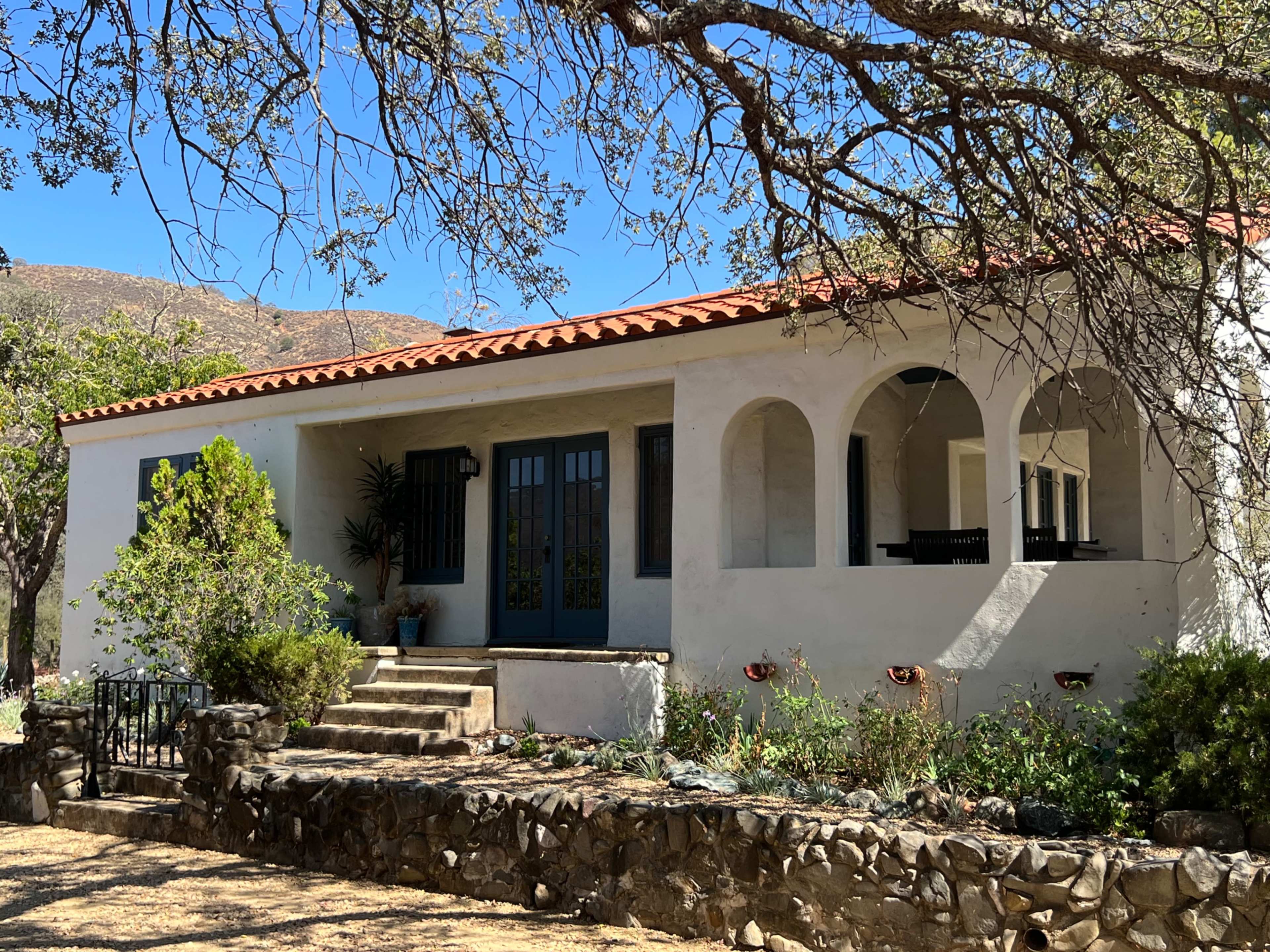 A white, single-story house with a red-tiled roof features arched doorways and a stone walkway surrounded by greenery.