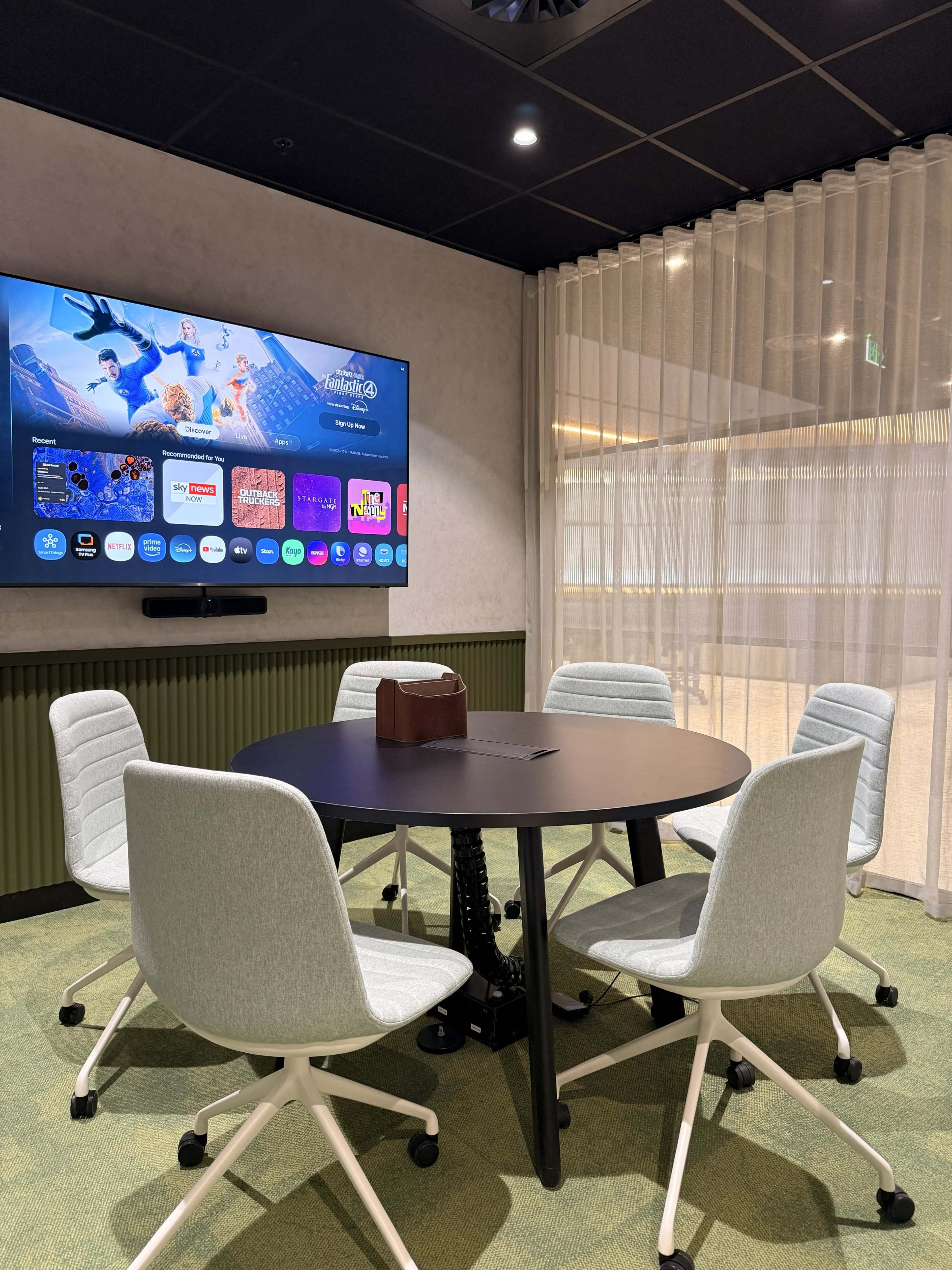 A modern meeting room features a circular table surrounded by four light gray chairs, with a large screen displaying various applications and content on the wall.