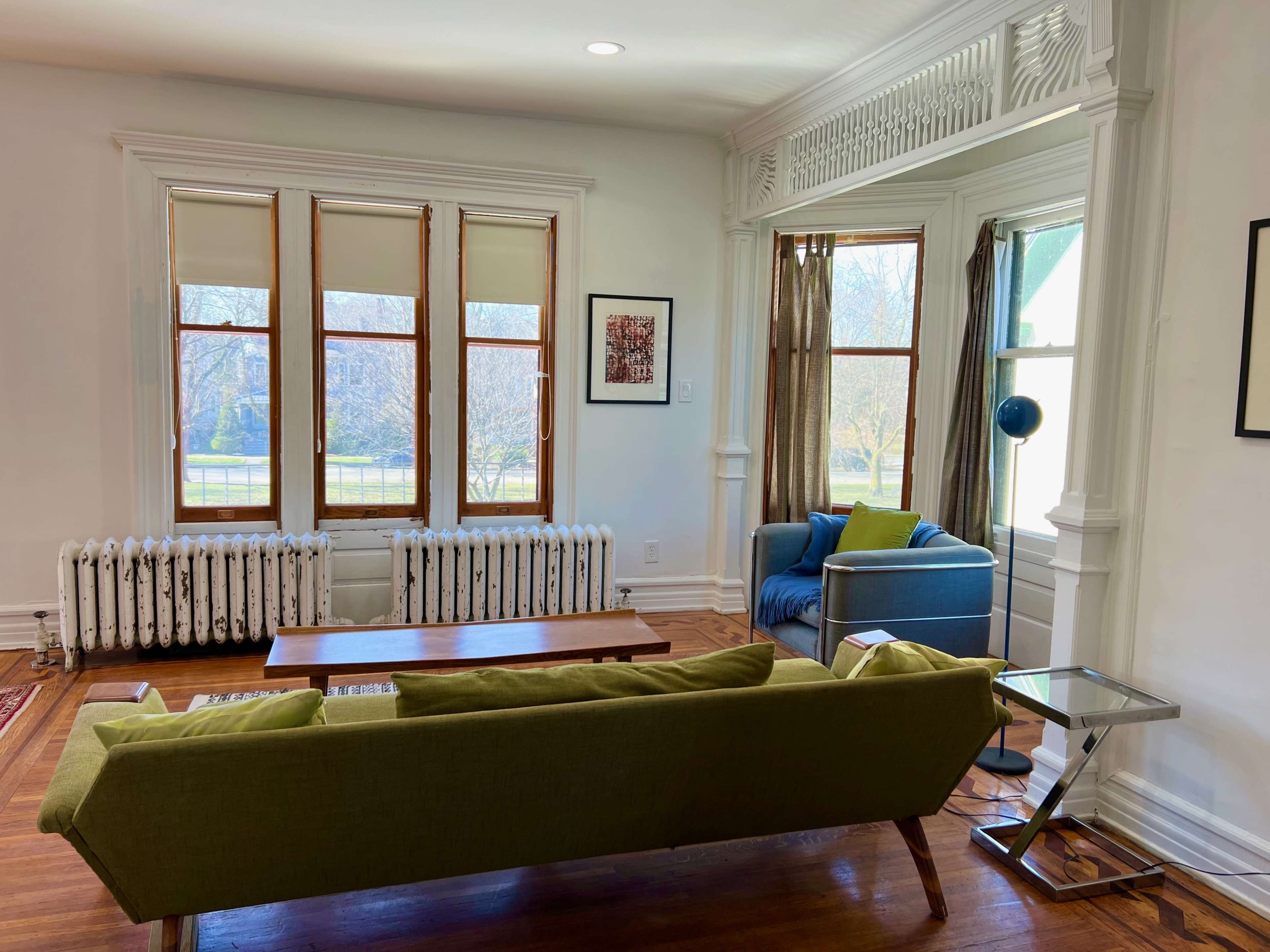 A bright living room with a green sofa, a blue armchair, a wooden coffee table, and large windows overlooking a garden.