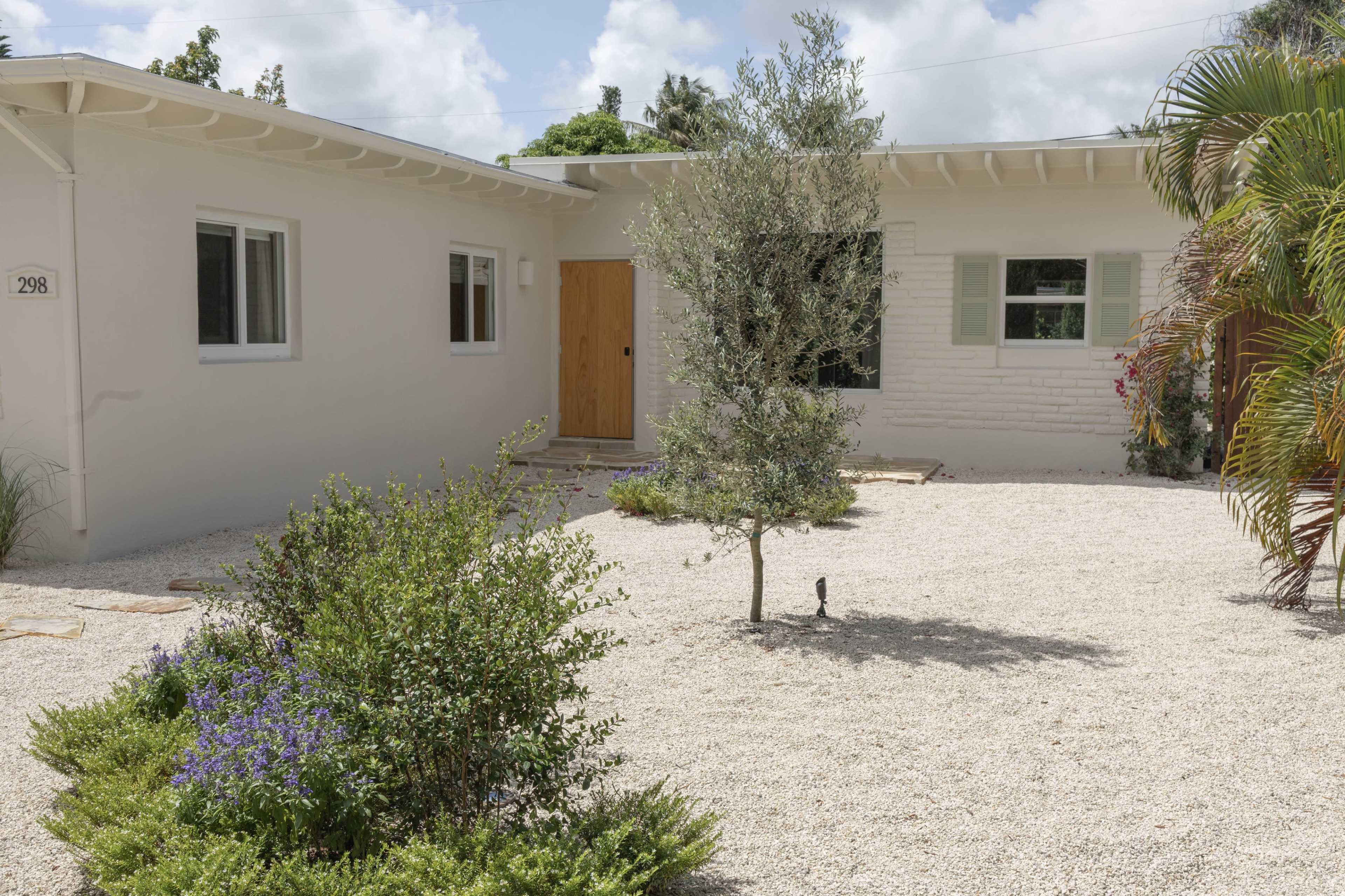 A small, one-story, light-colored house with a wooden door and windows is situated in a gravel yard featuring a small tree and purple flowering plants.