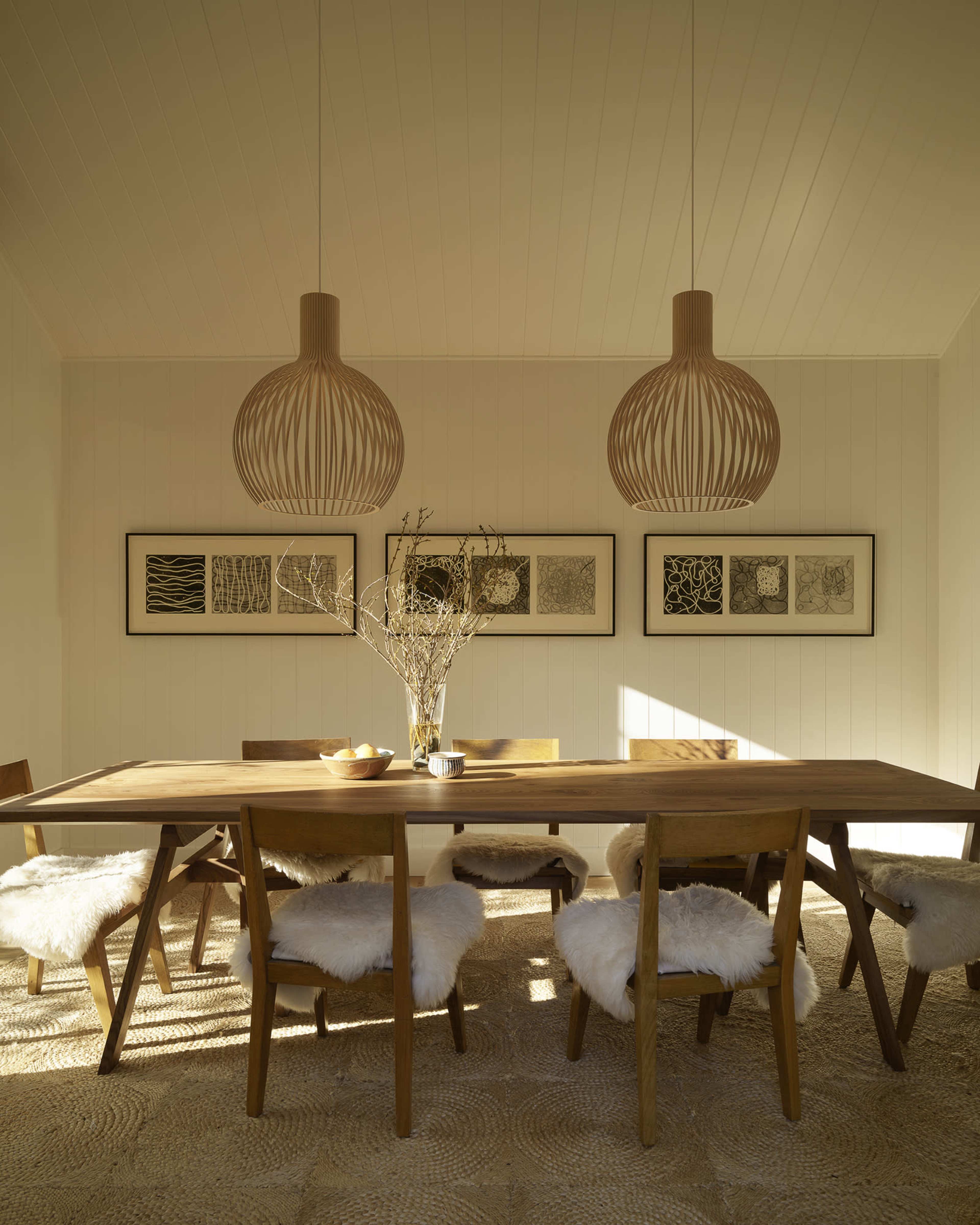 A bright dining room features a wooden table surrounded by chairs, with two pendant lights overhead and framed art on the walls.