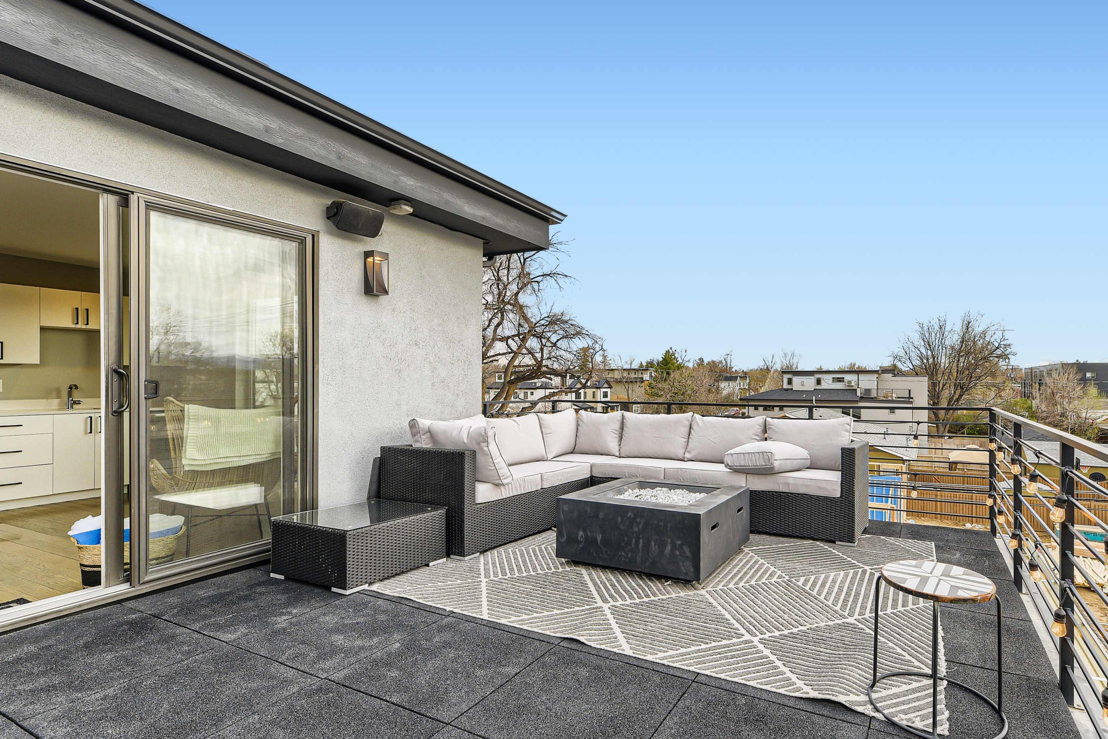 A modern rooftop terrace features a sectional sofa, a fire pit, and a small side table overlooking a neighborhood.