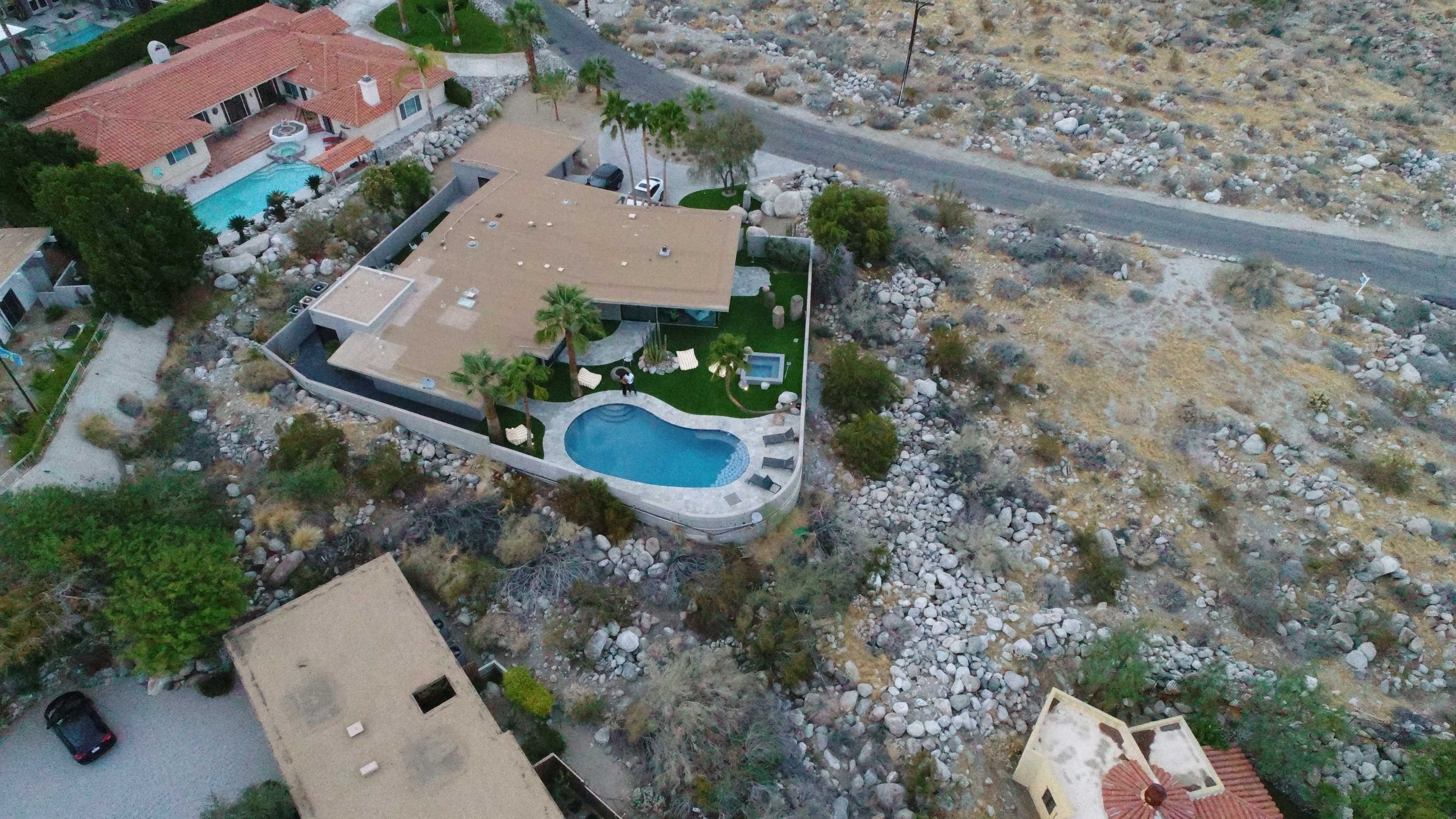 An aerial view shows a modern house with a swimming pool surrounded by rocky terrain and other residential buildings nearby.