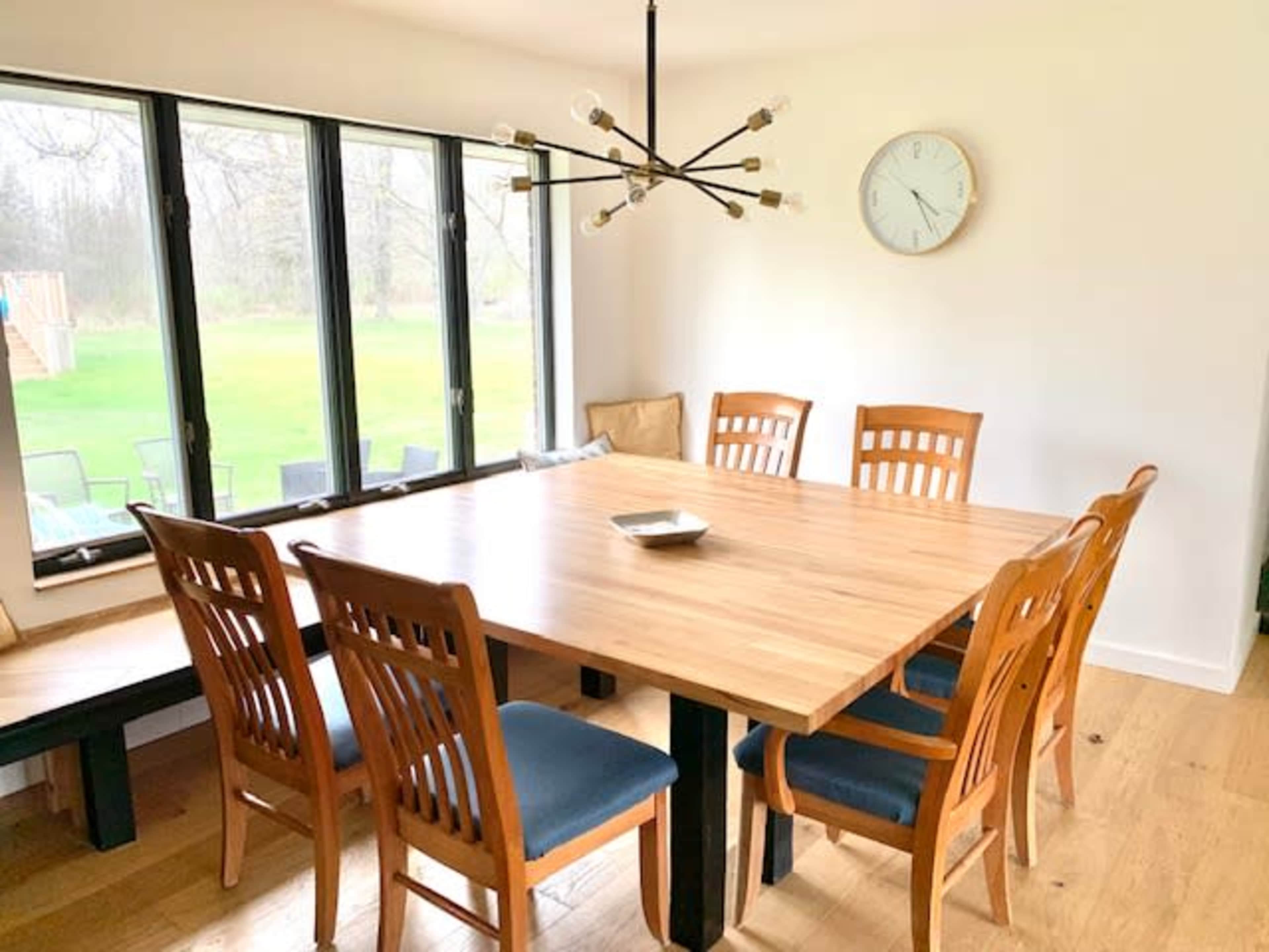 A wooden dining table with eight chairs is positioned near large windows that overlook a grassy outdoor area.