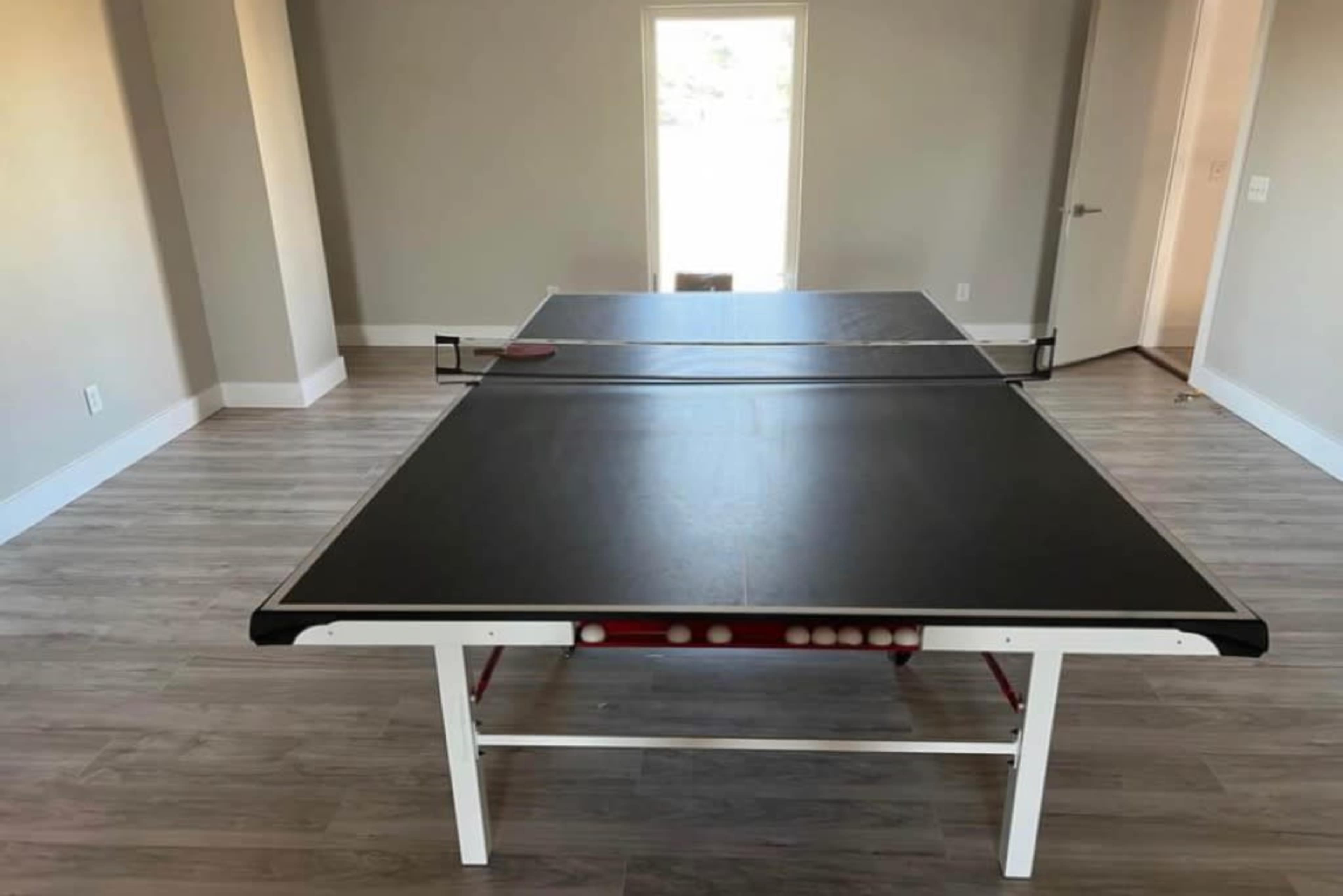 A ping pong table is positioned in a sparsely furnished room with light-colored walls and a window in the background.