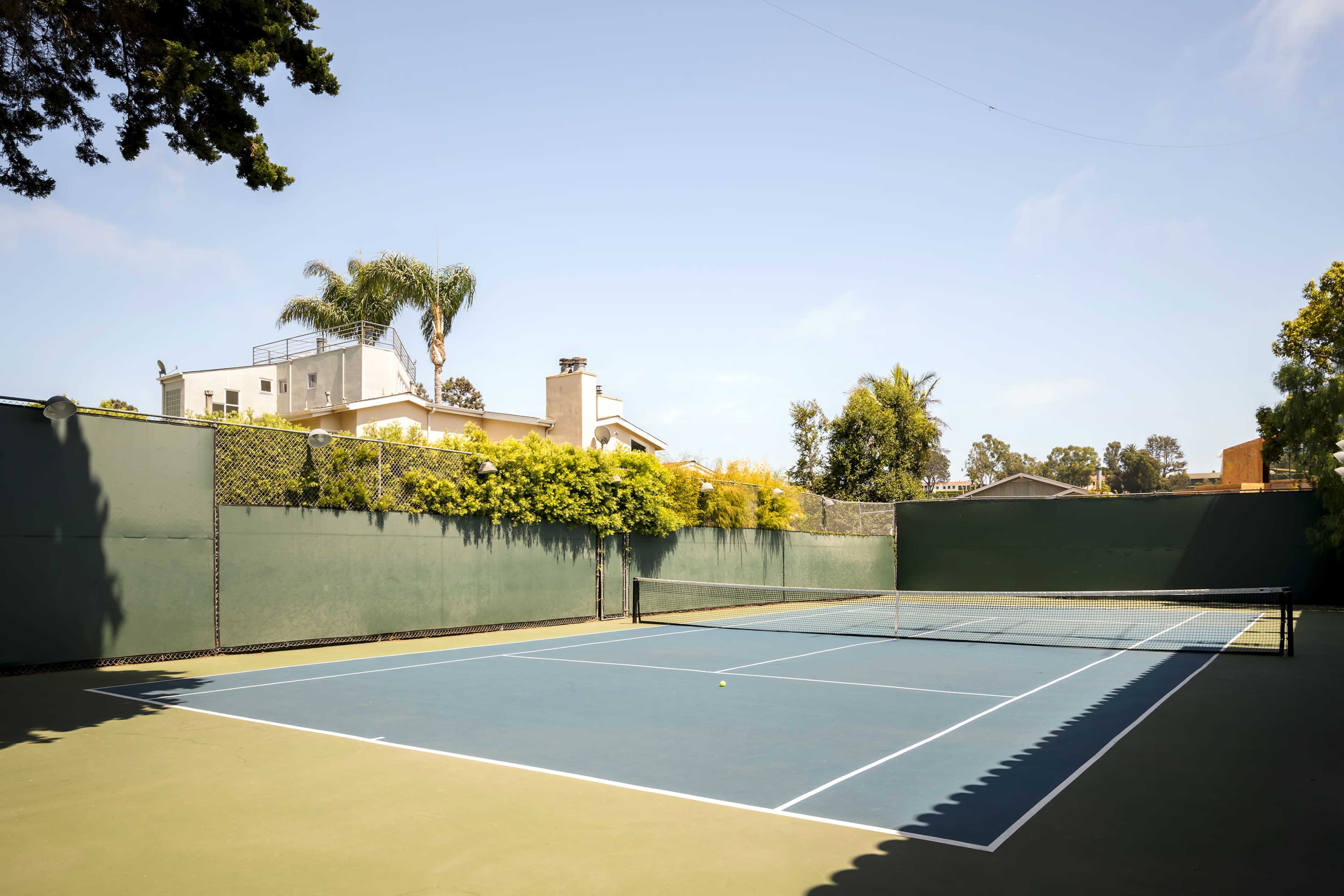 A tennis court with a blue surface and green surrounding walls is enclosed by trees and buildings in a residential area.